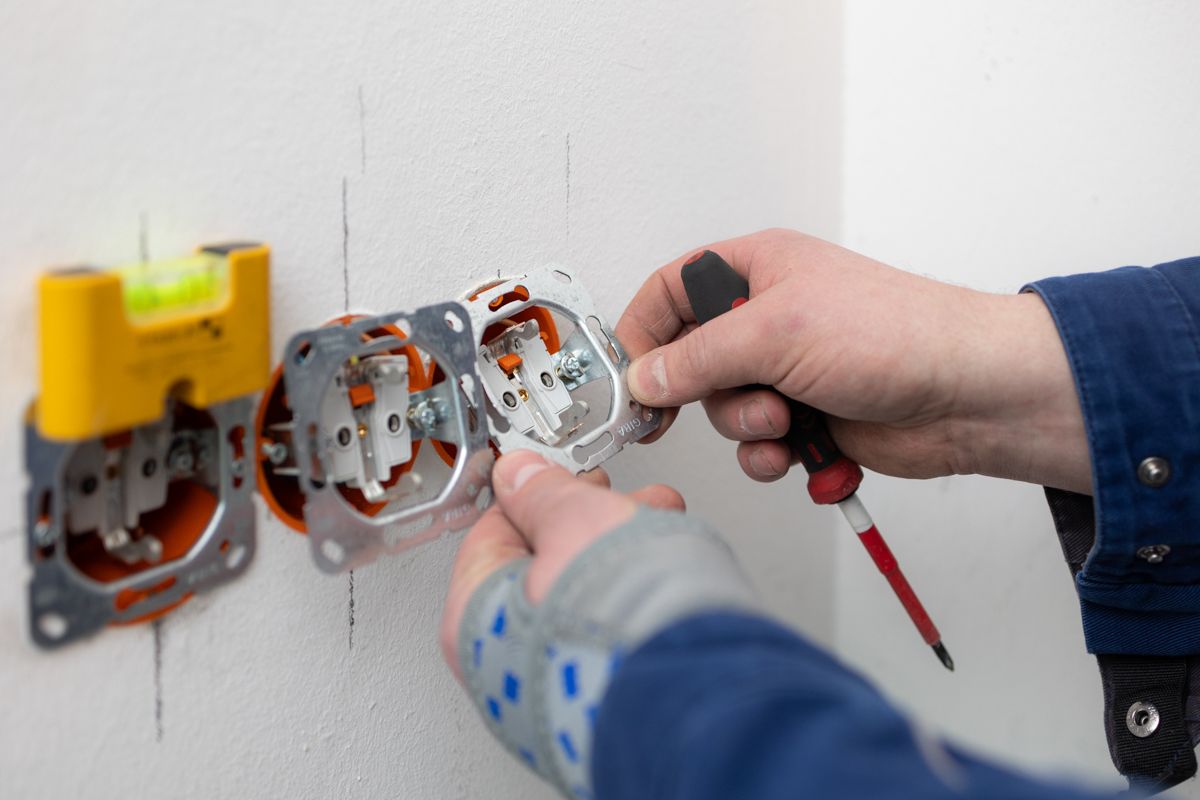 Hände verkabeln eine Steckdose in einer Wand, Dosen und Werkzeuge sind sichtbar.