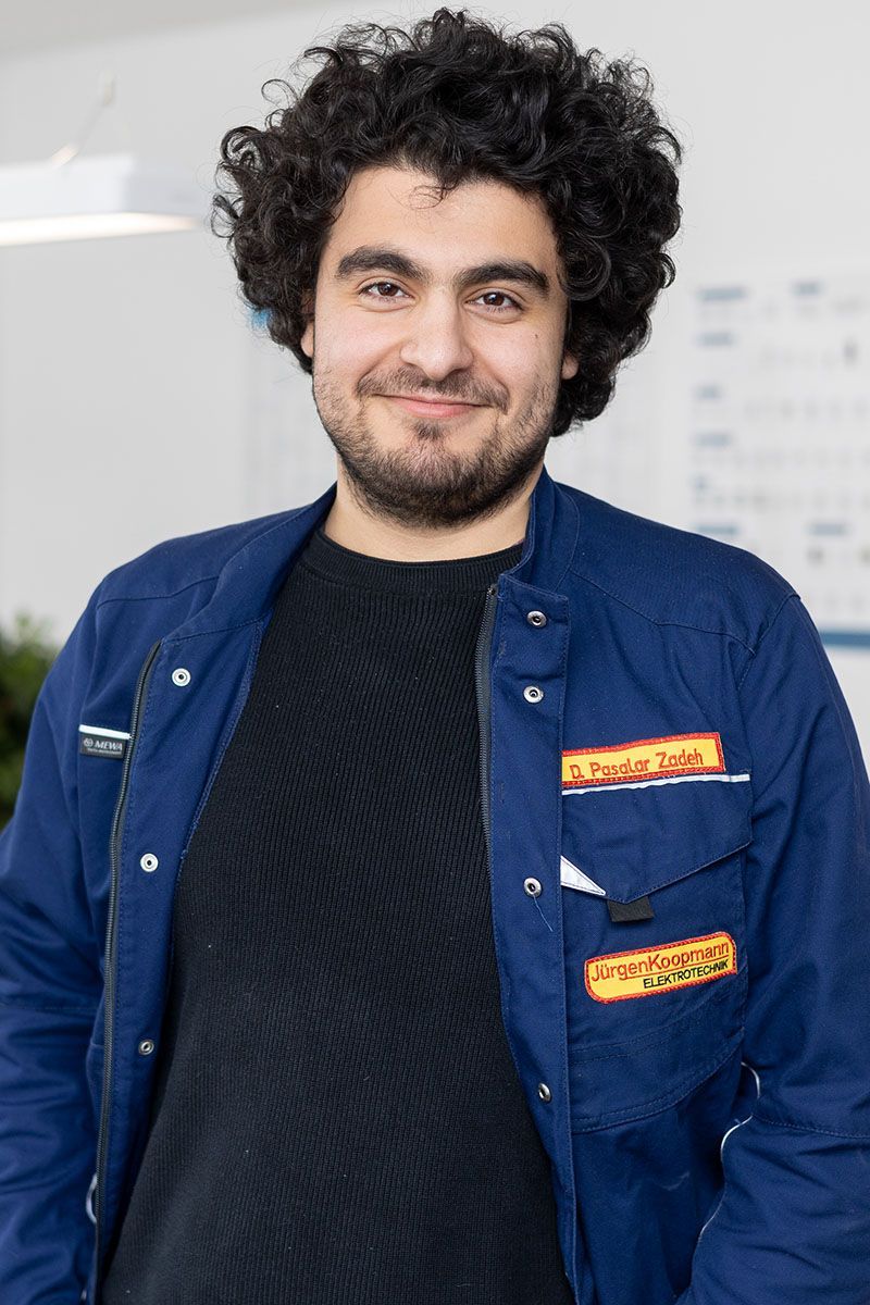 Ein Mann in einer blauen Jacke und einem schwarzen Hemd steht vor einem Tisch.