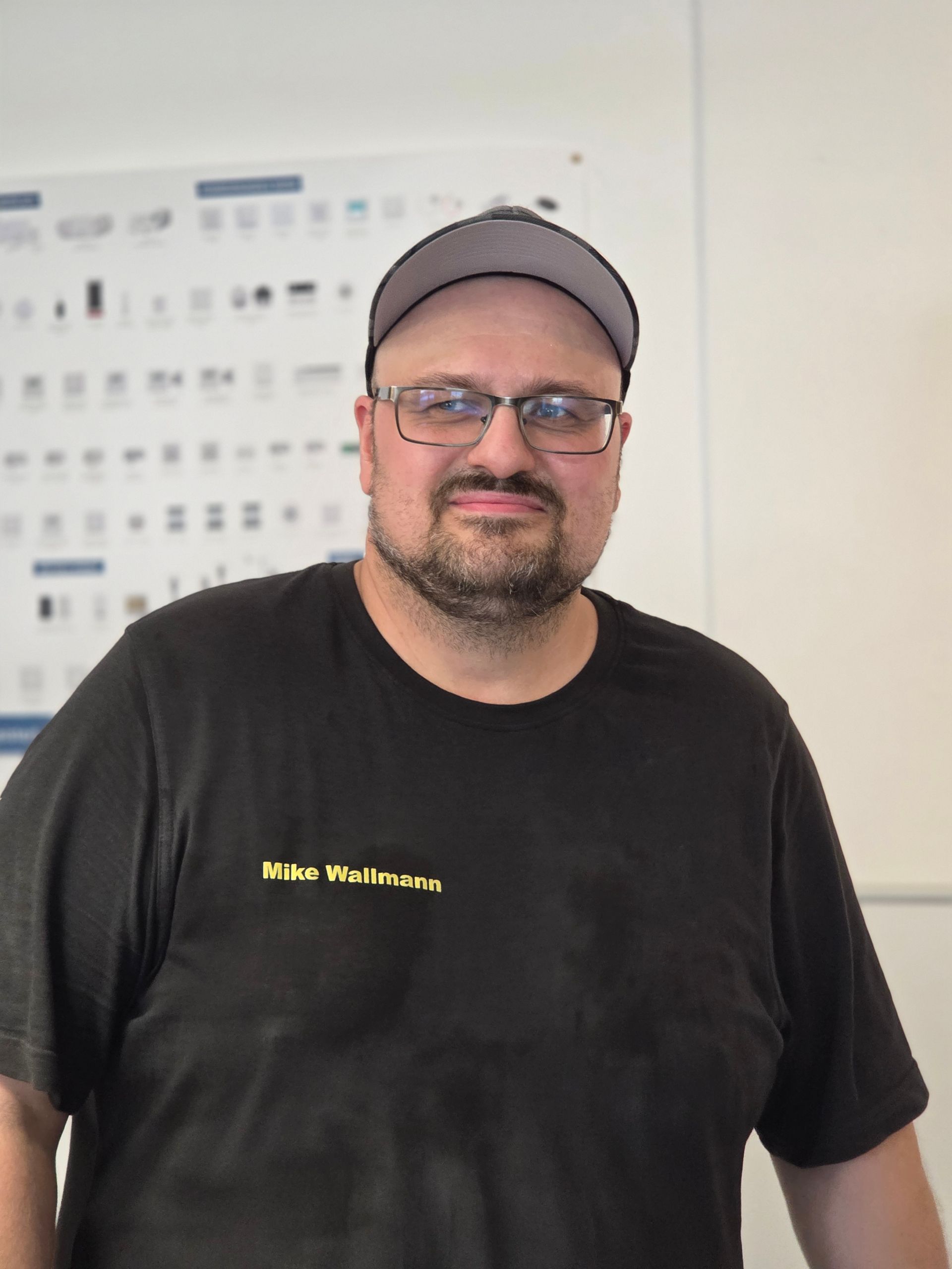 Mann mit grauer Kappe und Brille, bekleidet mit einem schwarzen T-Shirt, steht in einem Innenraum und blickt in die Kamera.