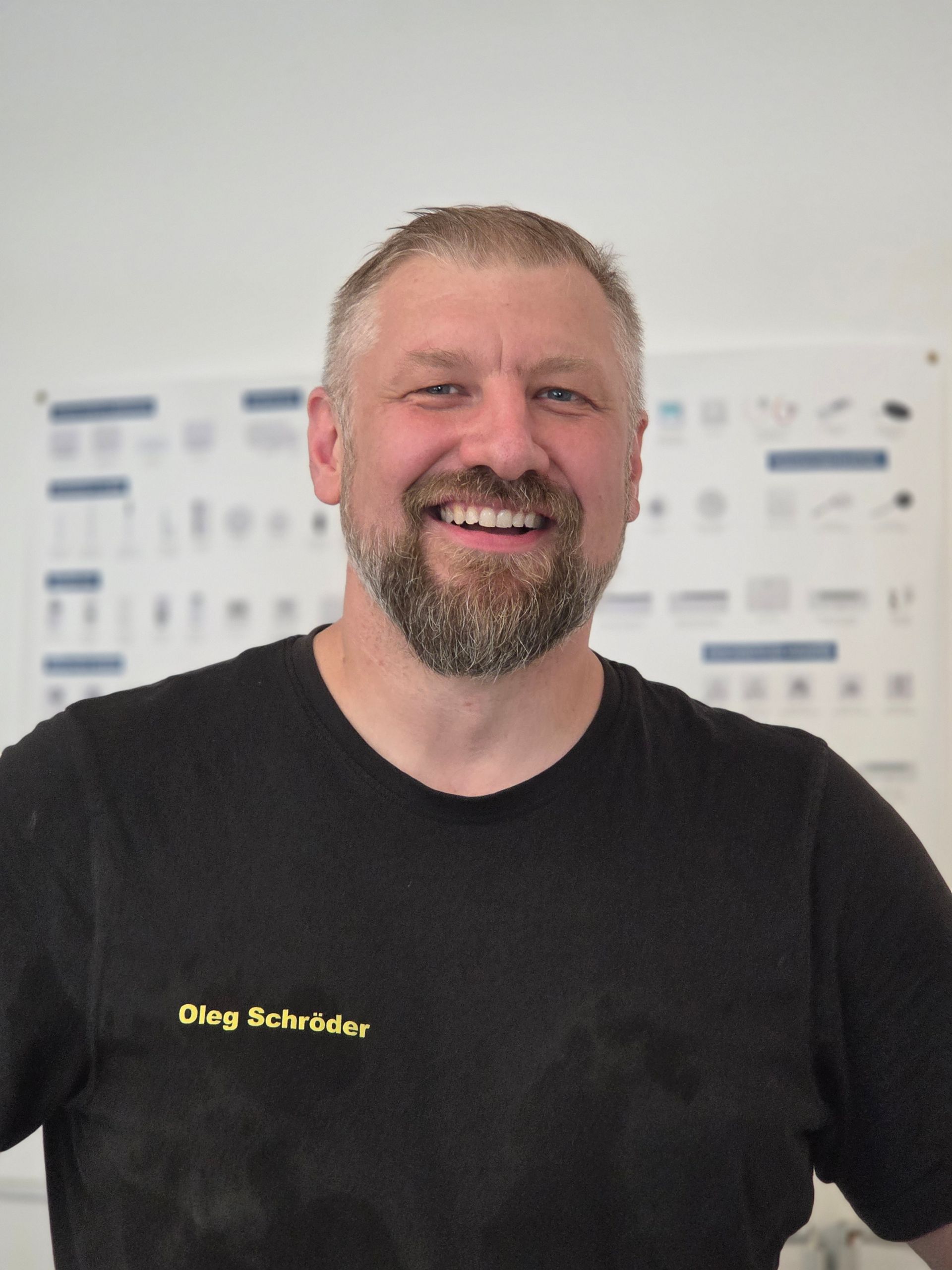 Ein lächelnder Mann in einem schwarzen T-Shirt steht in einem Innenraum vor einer verschwommenen Wand mit Diagrammen.