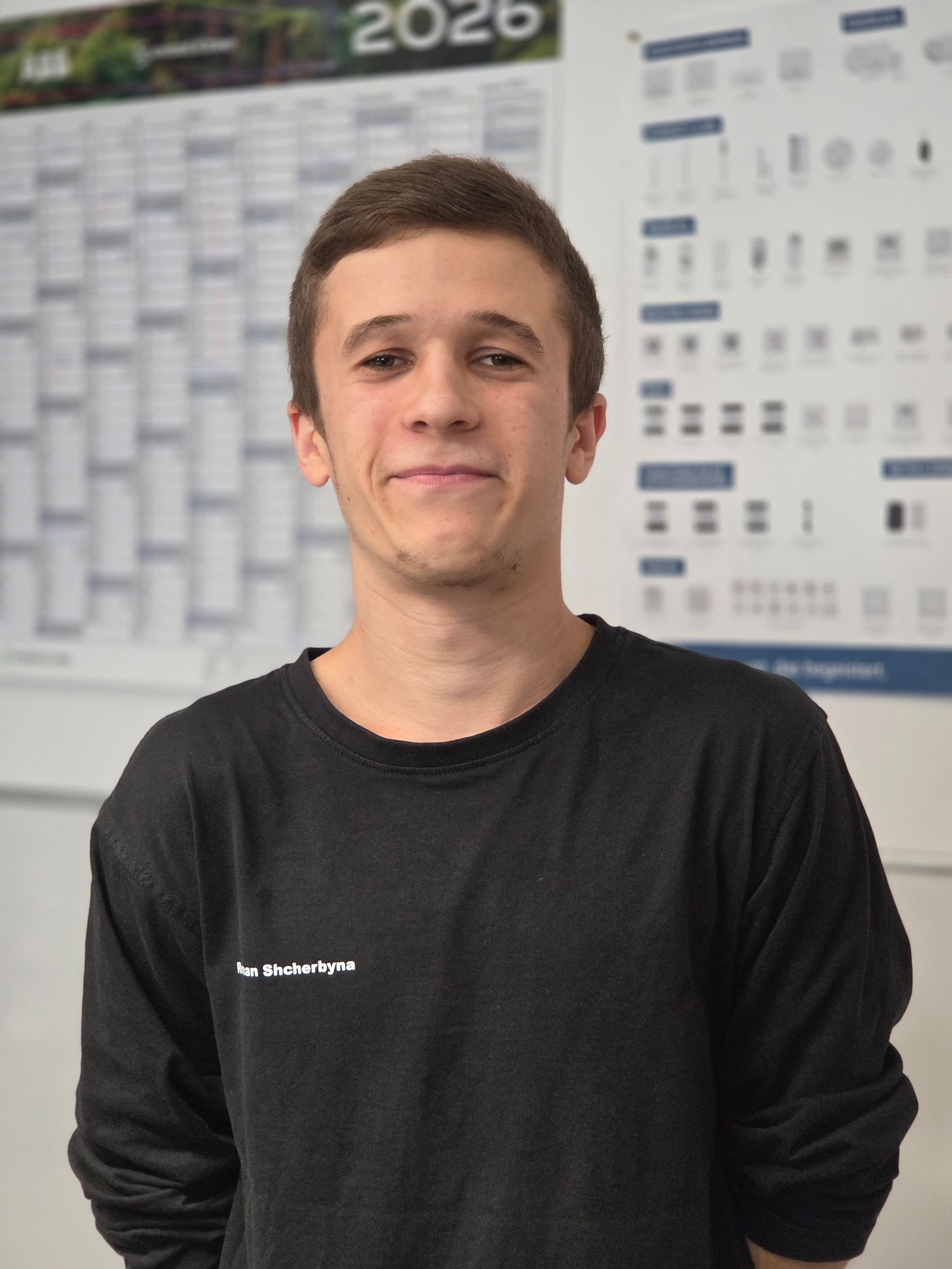 Eine Person steht vor einem Wandkalender, trägt ein schwarzes Hemd und lächelt leicht.
