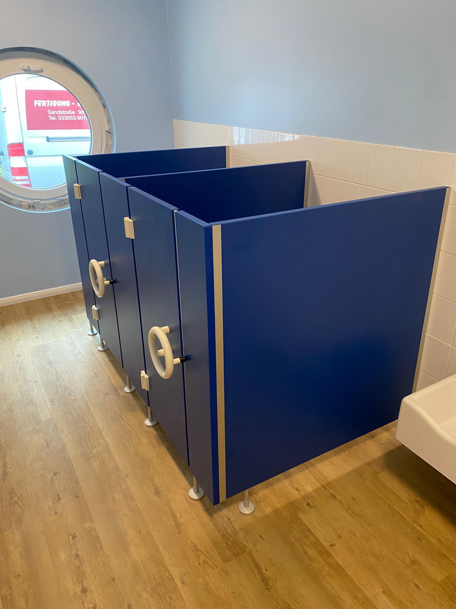 Ein Badezimmer mit blauen Trennwänden und einem runden Fenster.