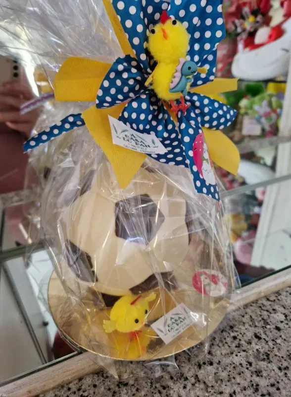Balón de fútbol de chocolate envuelto en celofán, decorado con lazos amarillos y azules y pequeños pollitos de juguete.