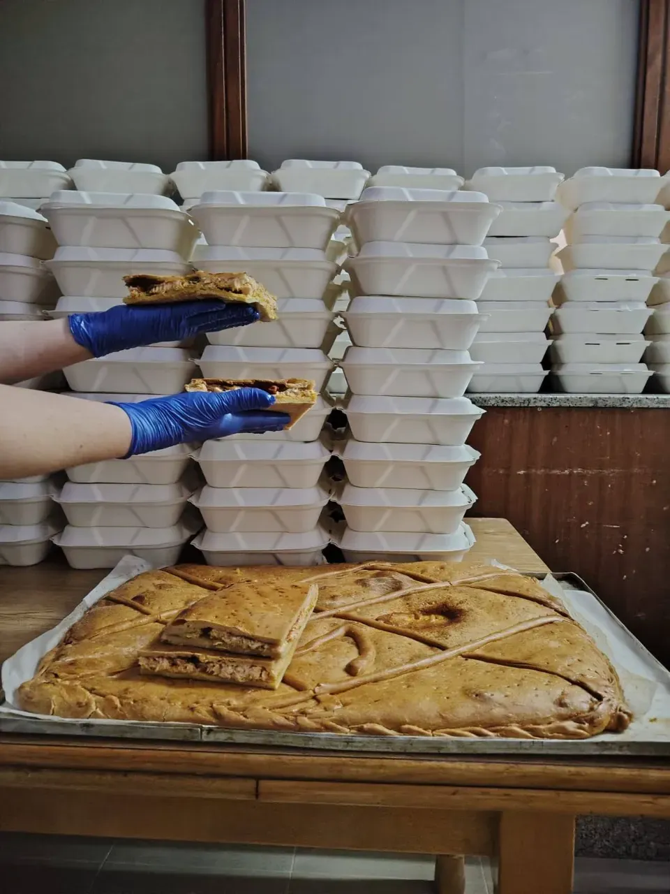 Manos con guantes azules sosteniendo rebanadas de empanada, una grande en una bandeja y pilas de recipientes para llevar.