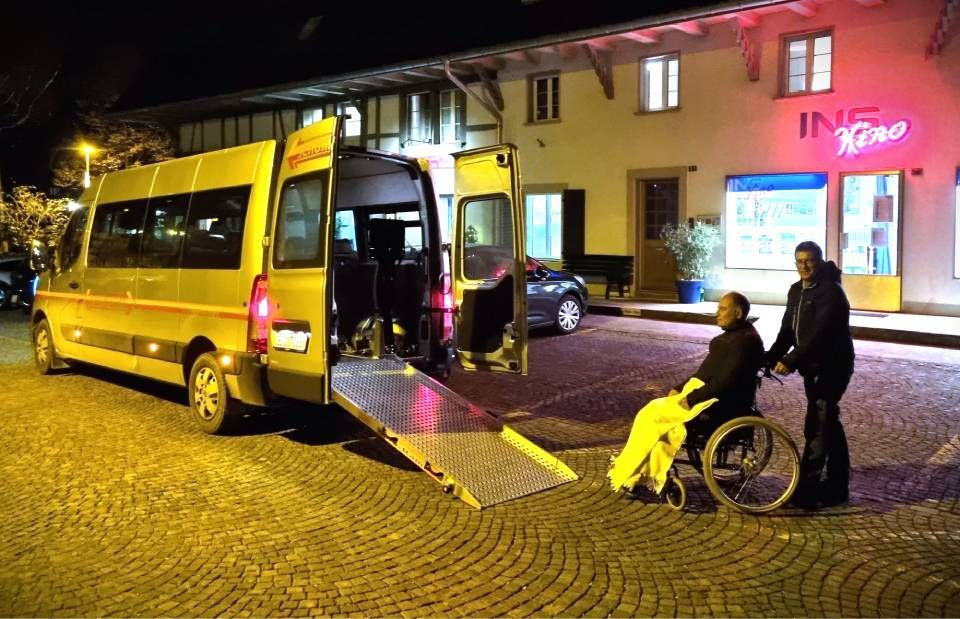 Gelber Kleinbus mit ausgefahrener Rampe bei Nacht, ein Rollstuhlfahrer steigt neben zwei Helfern ein.
