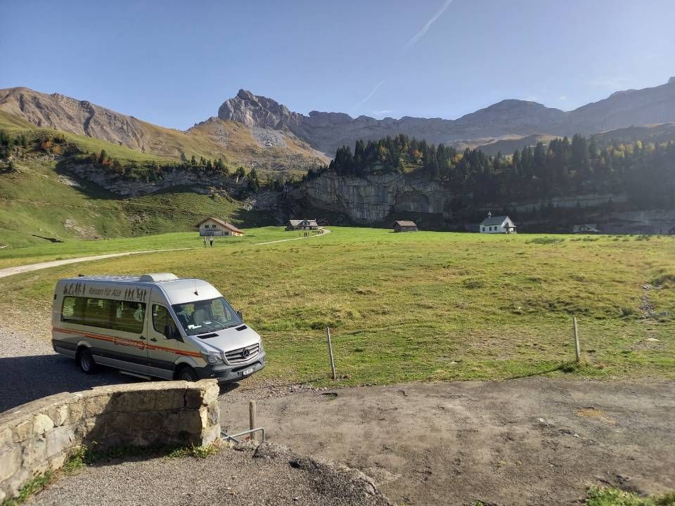 Ein grüner Lieferwagen parkt neben einer Bergwiese mit felsigen Hügeln und Hütten im Hintergrund.