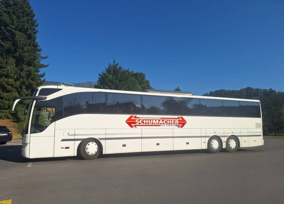 Ein weisser Reisebus mit rotem Logo parkt auf einem gepflasterten Parkplatz unter klarem, blauem Himmel.
