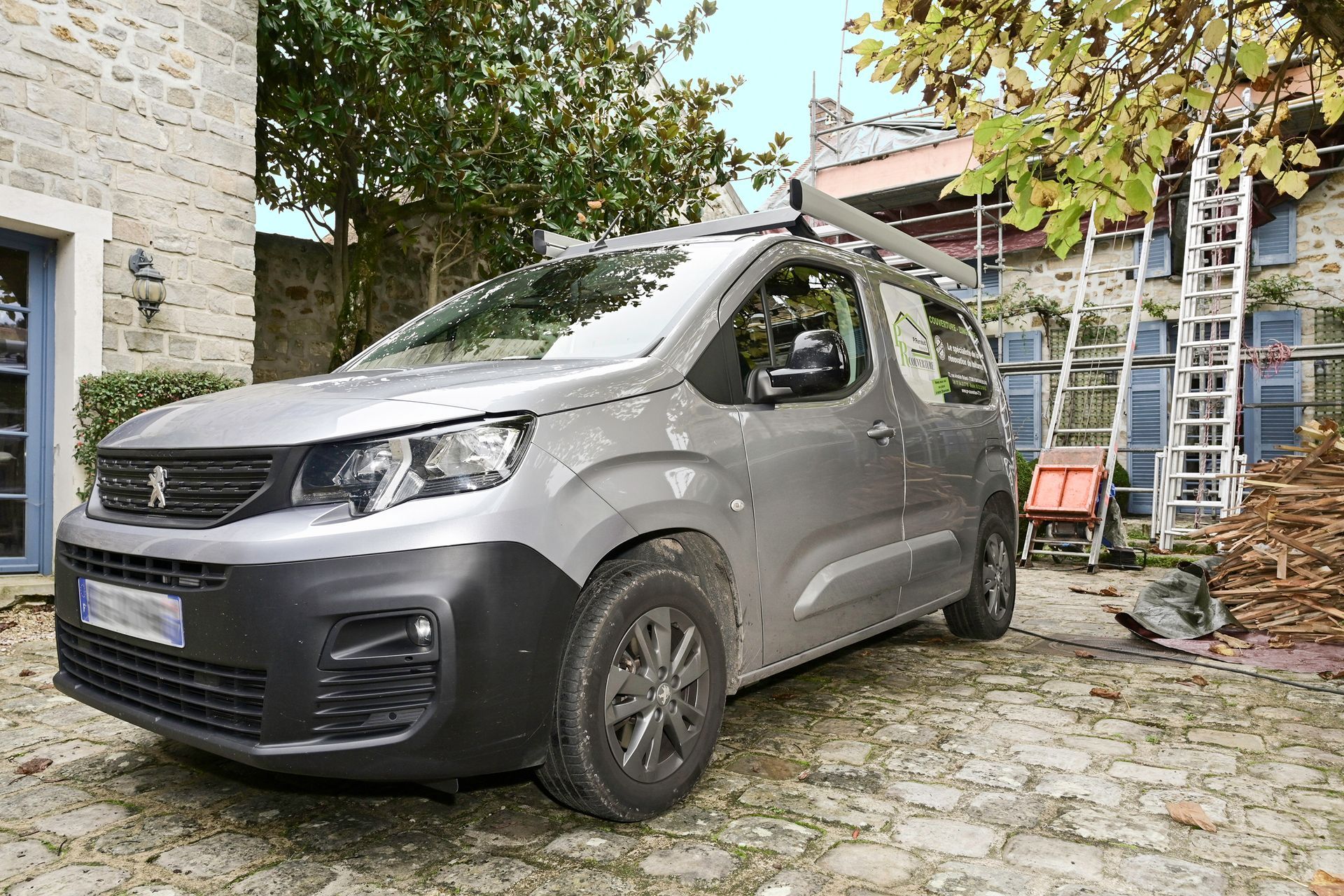 Véhicule utilitaire gris garé dans une cour pavée entouré d'outils avec une maison en pierre et des arbres en arrière-plan