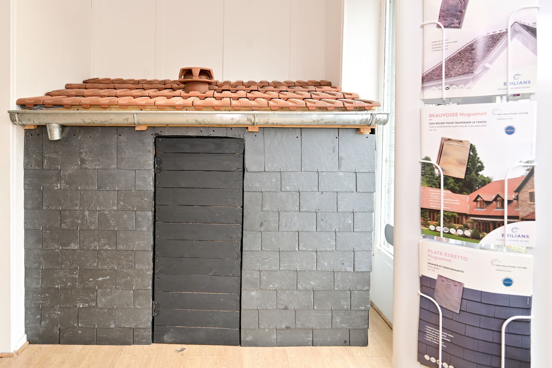 Maquette de toiture avec tuiles et ardoises exposée à l'intérieur, accompagnée de panneaux d'information