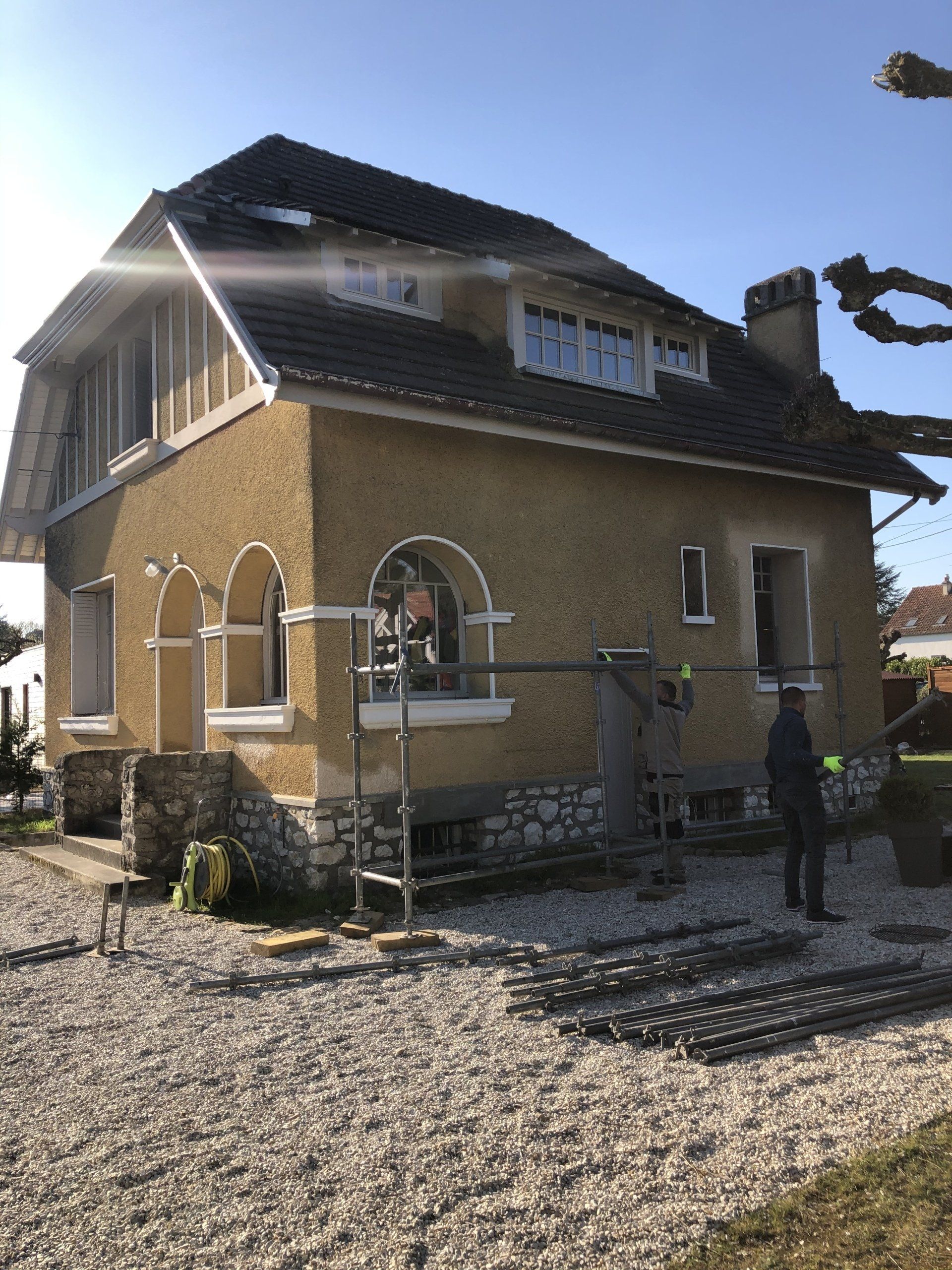 Maison en crépi beige avec toit en rénovation et artisan travaillant sur la droite