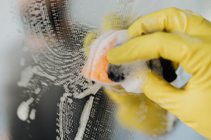 Main gantée de jaune frottant une surface vitrée avec une éponge savonneuse, mousse nettoyante visible