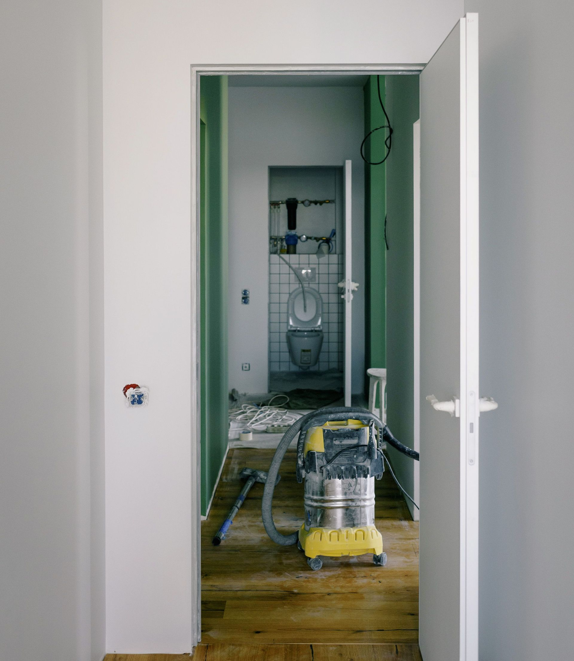 Couloir en rénovation, avec des outils et un aspirateur au sol et une porte donnant sur une salle de bains en construction.