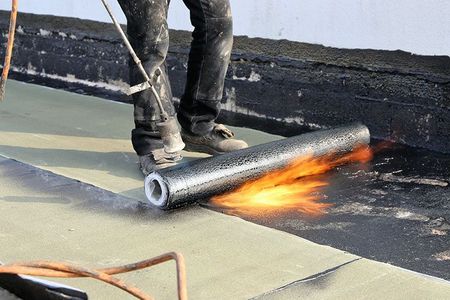 Eine Person rollt mit einem Brenner eine Rolle Dachmaterial auf ein Dach.