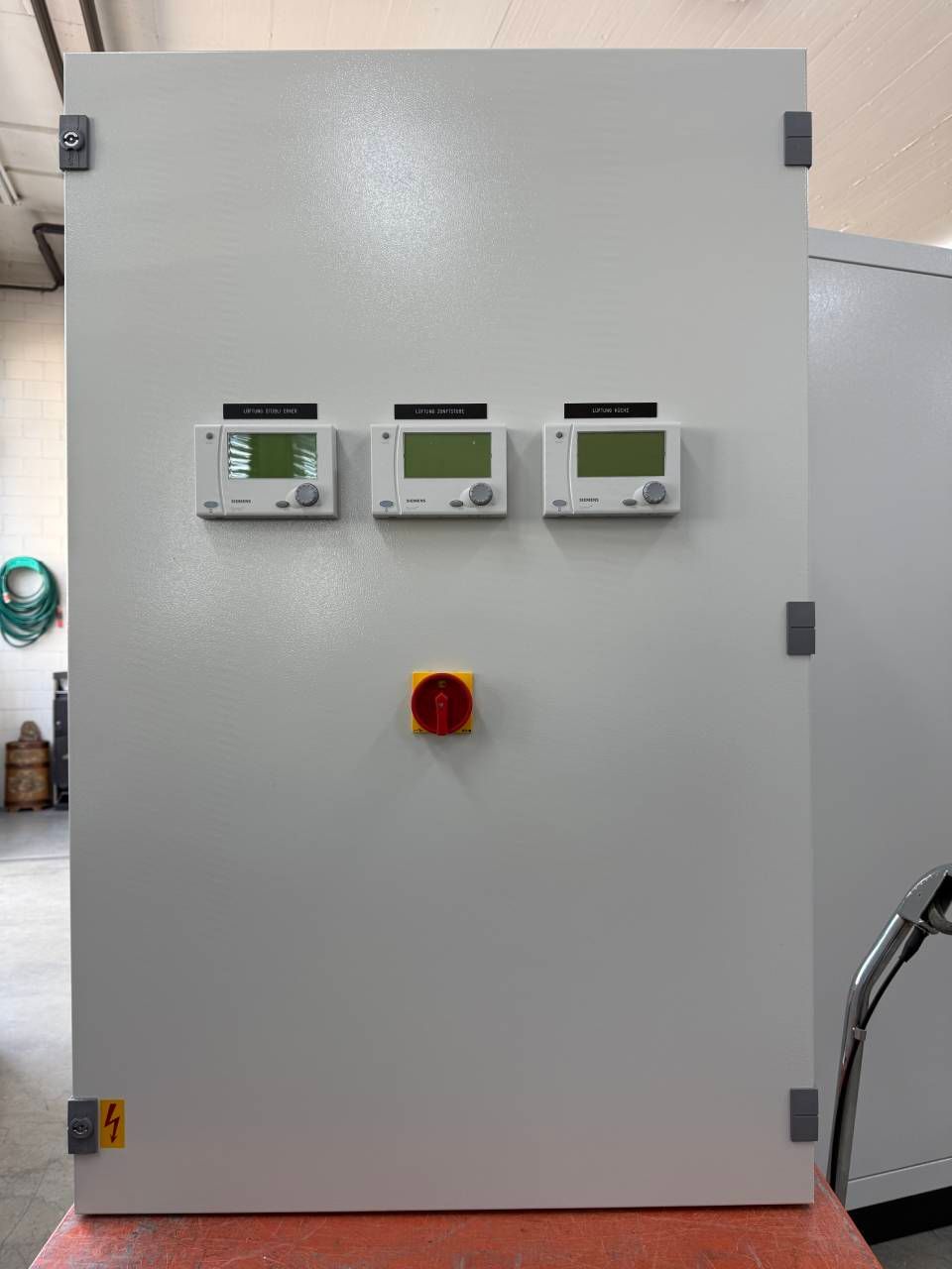 Graues elektrisches Bedienfeld mit drei kleinen Anzeigen und einem roten Not-Aus-Knopf.