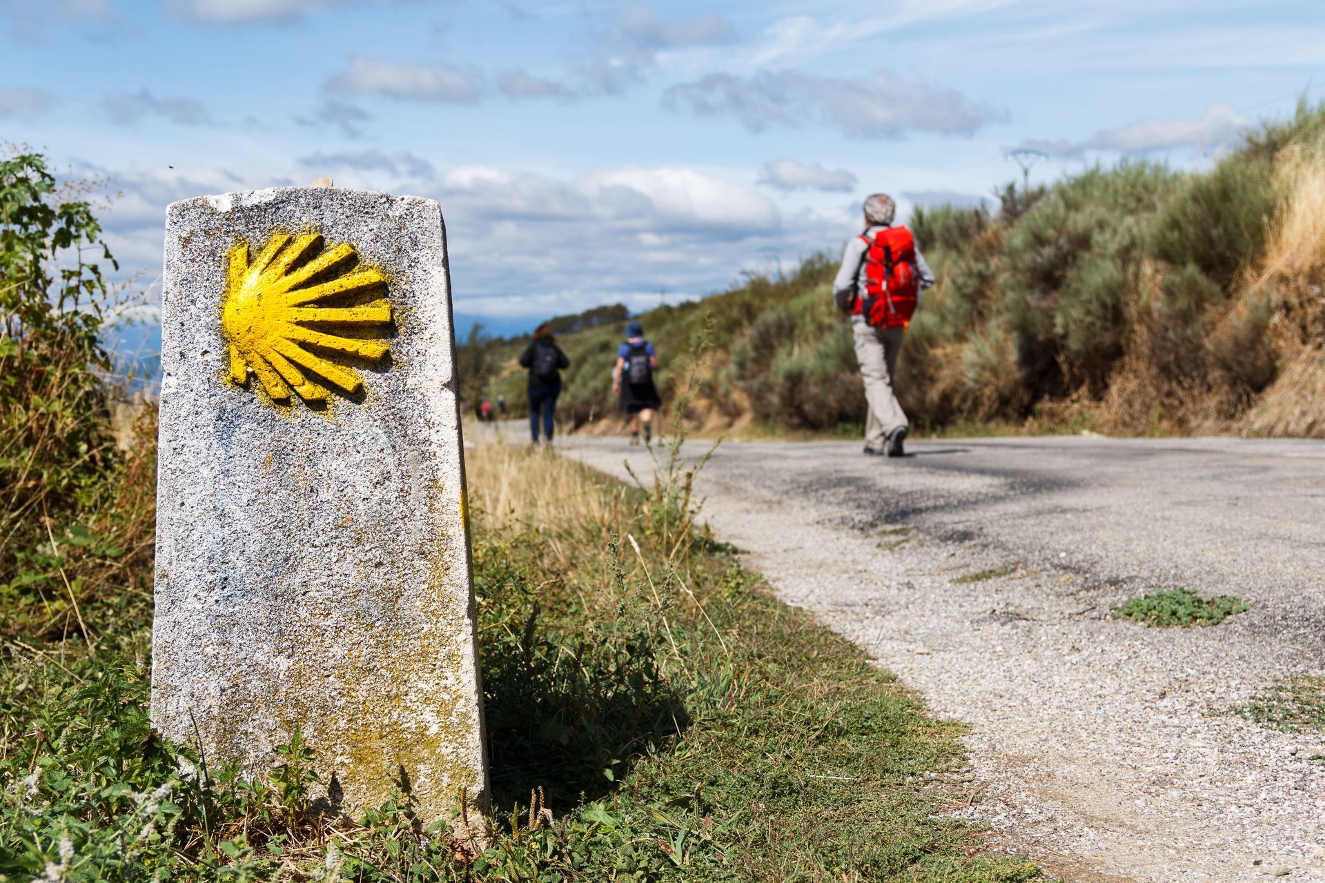 Randonneurs sur un chemin de campagne ensoleillé, à côté d'une borne kilométrique en pierre ornée d'un symbole de coquillage jaune.