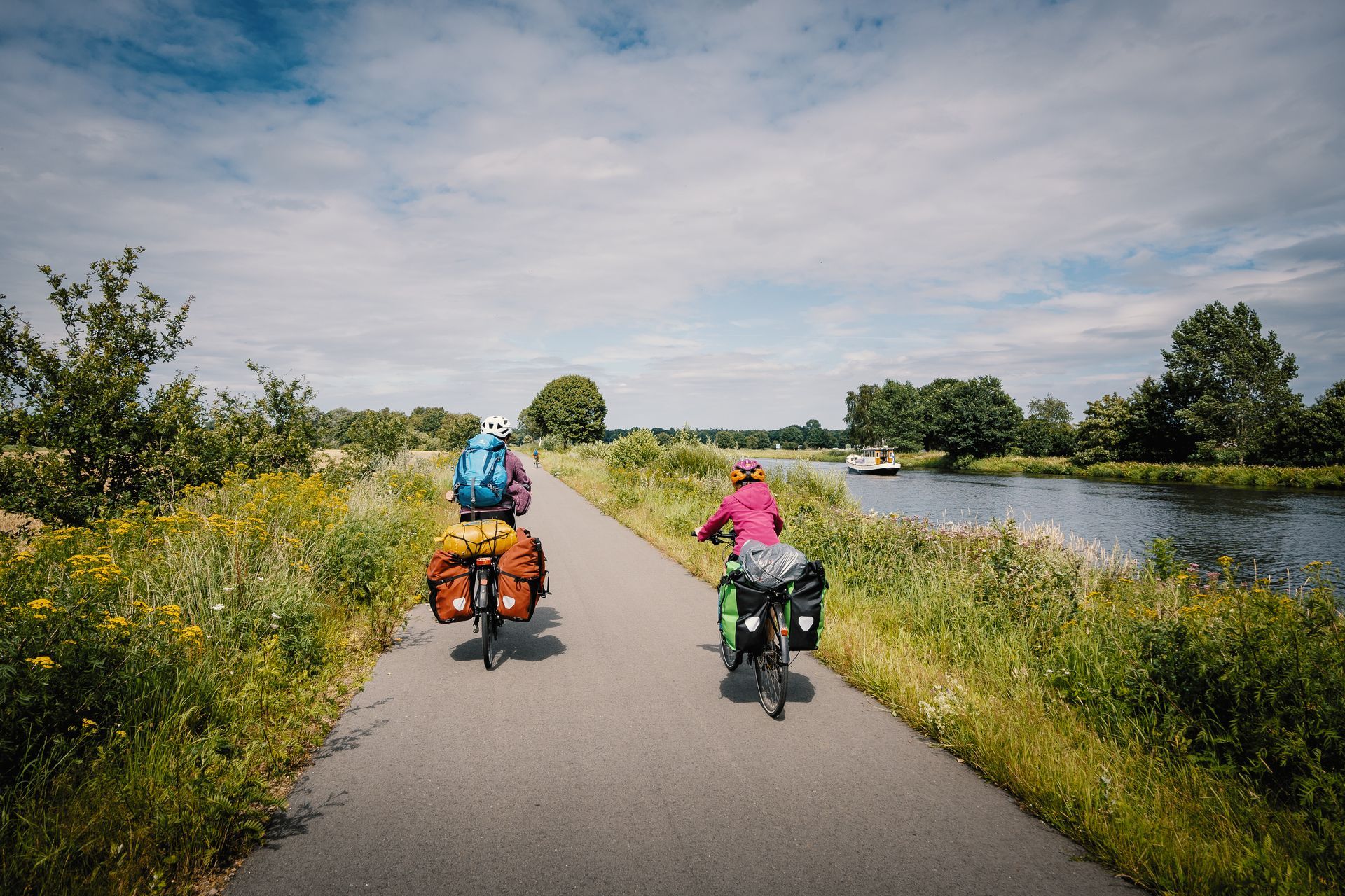 Deux cyclistes roulent sur une piste cyclable goudronnée longeant une rivière sous un ciel partiellement nuageux.