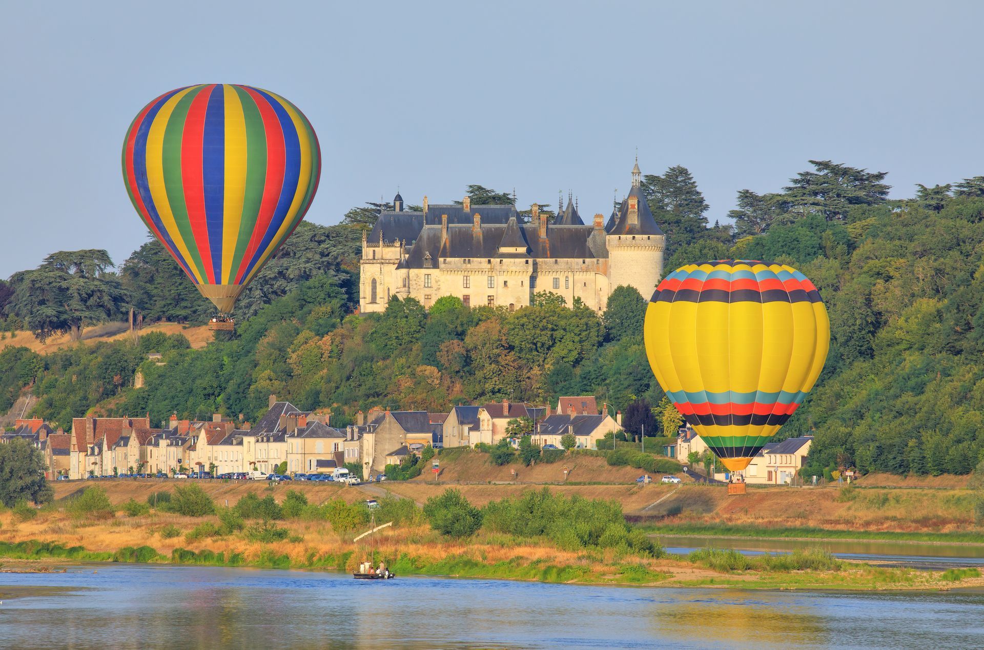 Deux montgolfières colorées flottent au-dessus d'un château et d'un village au bord de la rivière au lever du soleil.