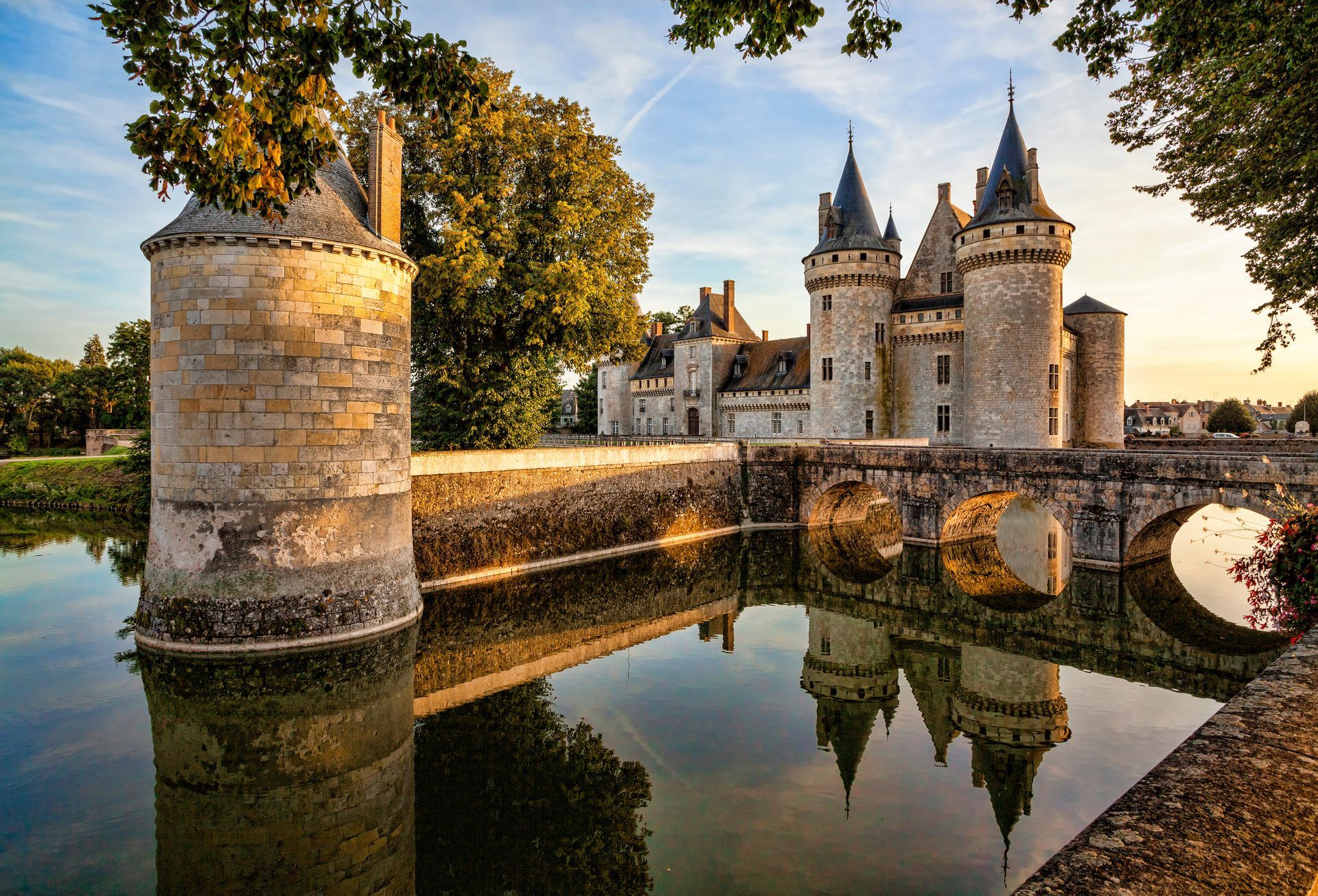 Château de pierre avec tours et douves au coucher du soleil, se reflétant dans l'eau calme près d'un pont.