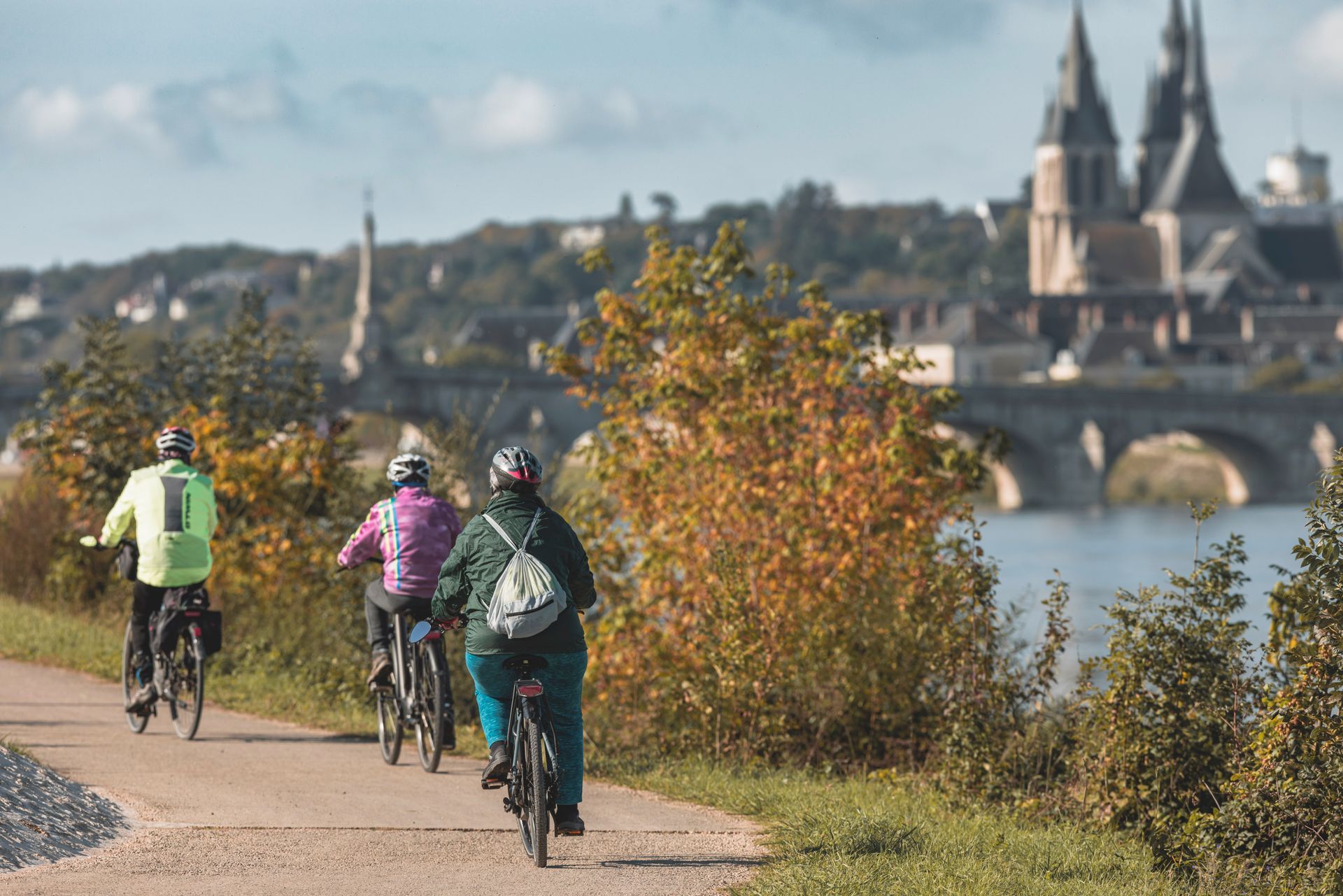 Des cyclistes roulent sur un chemin au bord de la rivière, bordé d'arbres aux couleurs de l'automne, avec un pont et un château en arrière-plan.