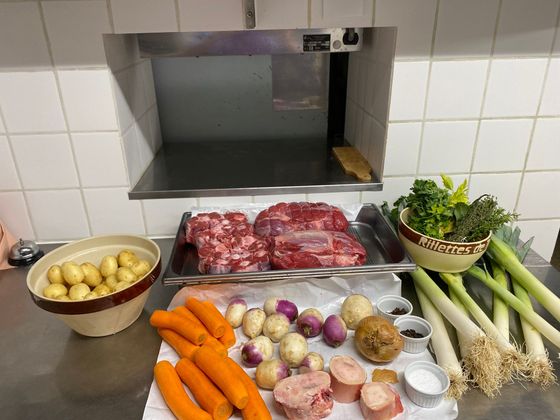 Ingrédients disposés sur un plan de travail, notamment de la viande, des pommes de terre, des carottes, des oignons, des poireaux, des légumes verts et des herbes aromatiques.