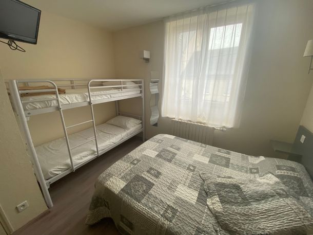 Petite chambre avec lits superposés, un lit double, une télévision murale et une fenêtre avec des voilages.