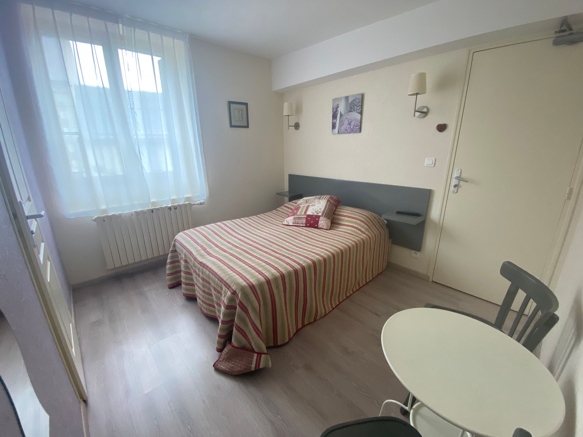 Petite chambre avec un lit simple, une fenêtre avec des voilages, une table ronde et une chaise.