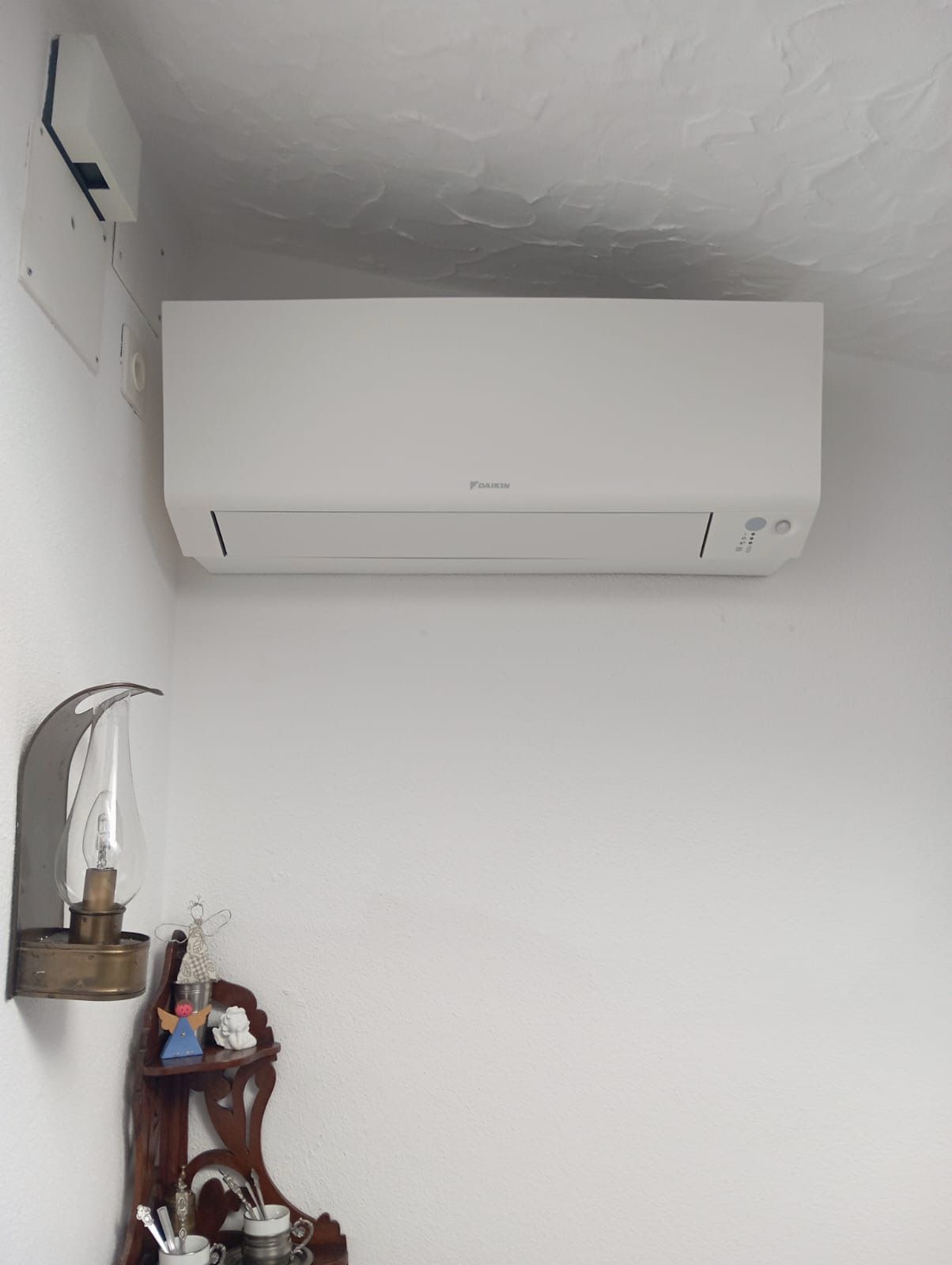 Condizionatore bianco su una parete bianca; lampada e mensola decorativa sotto.