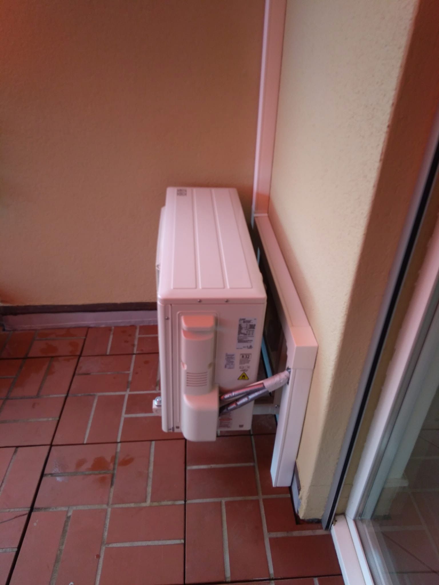 Condizionatore d'aria bianco installato su un patio piastrellato in mattoni, accanto a un muro e a una finestra color crema.