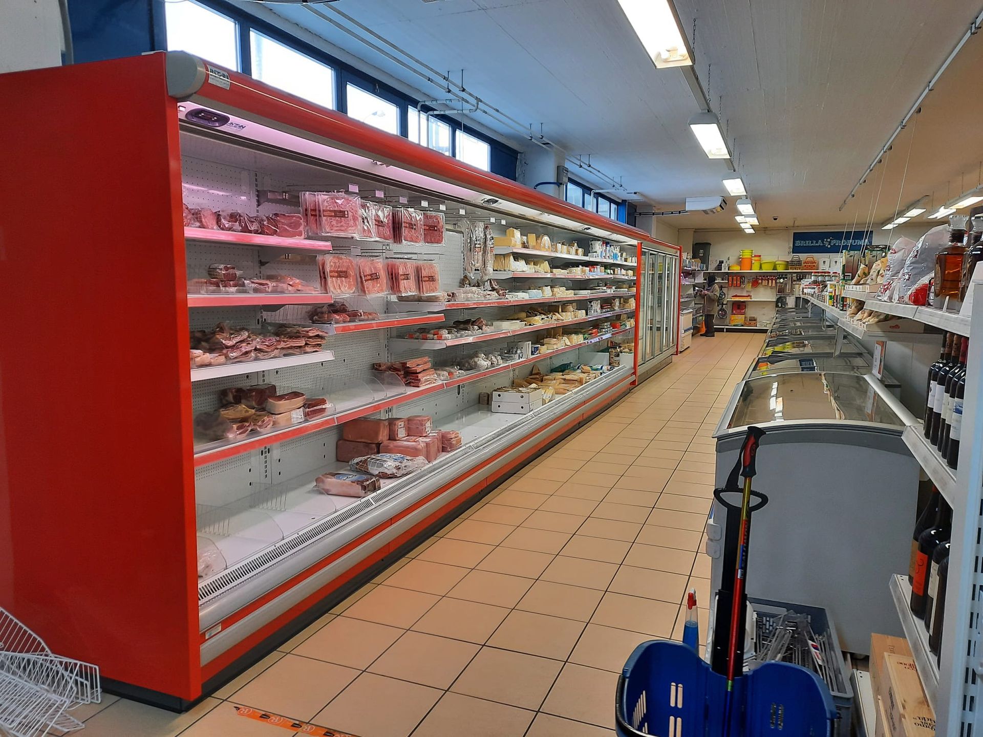 Una corsia di un supermercato. Una lunga vetrina refrigerata rossa espone salumi e formaggi.