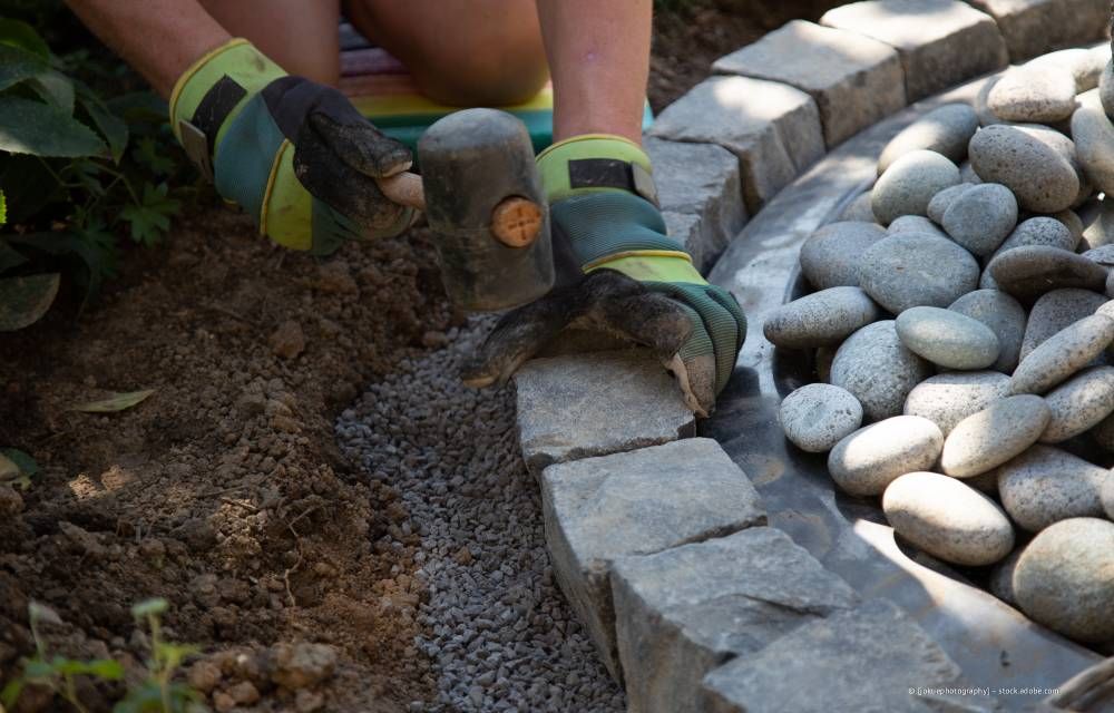 Behandschuhte Hände platzieren gebogene Steineinfassungen neben einem Kiesweg und dekorativen Flusssteinen.