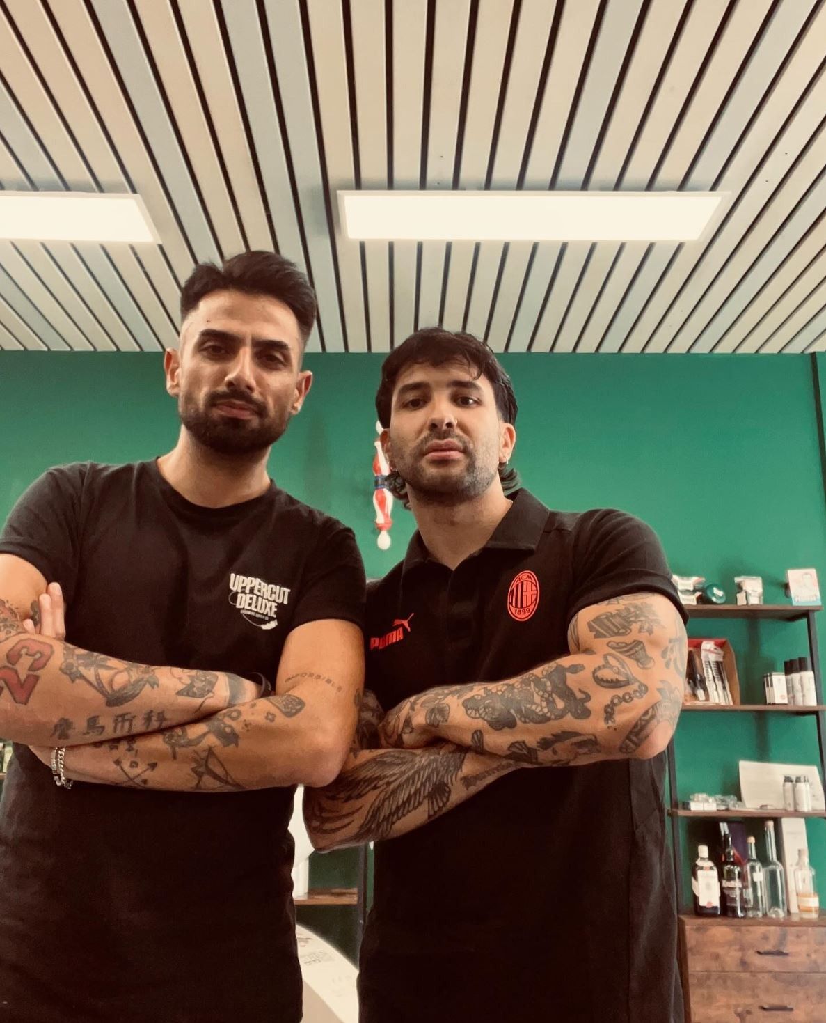 Due uomini tatuati sono in piedi in una stanza, con le braccia incrociate. Uno indossa una maglietta nera, l'altro una polo.