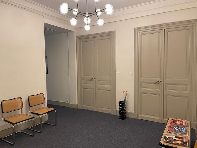 Pièce vide aux murs beiges, moquette bleue, chaises et portes doubles fermées sous un plafonnier