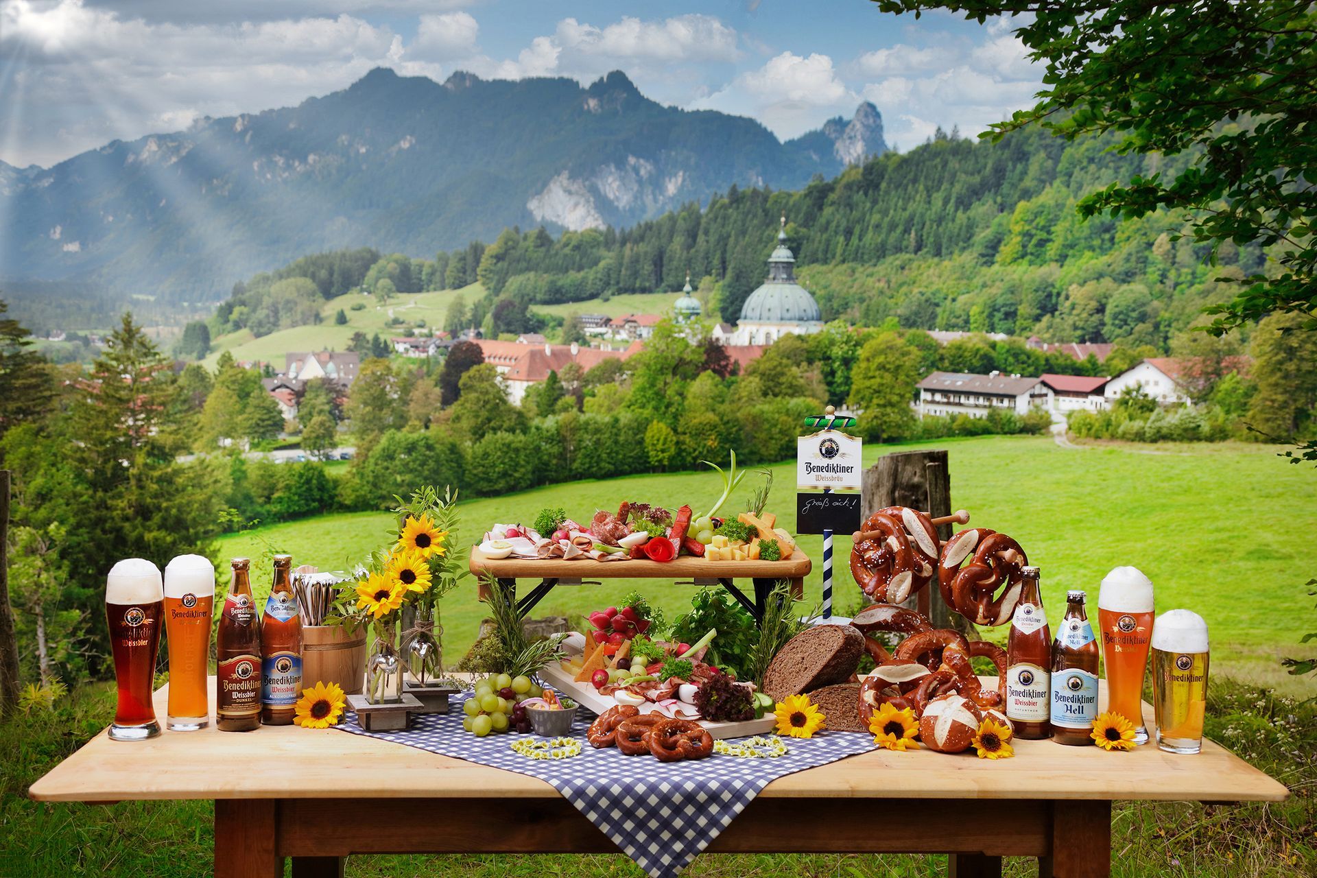 Bayerischer Tisch mit Brezn, Bier und regionalen Speisen vor einer Berglandschaft.