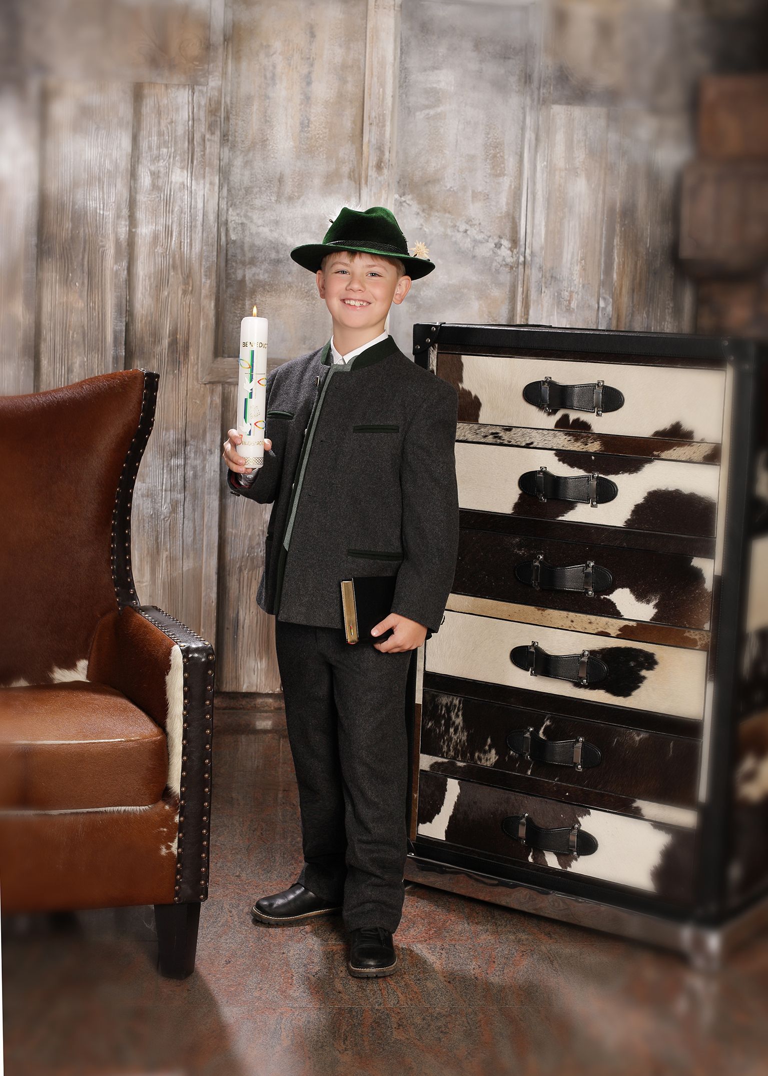 Junge in Tracht hält eine festlich geschmückte Kerze und sitzt vor rustikalem Hintergrund.