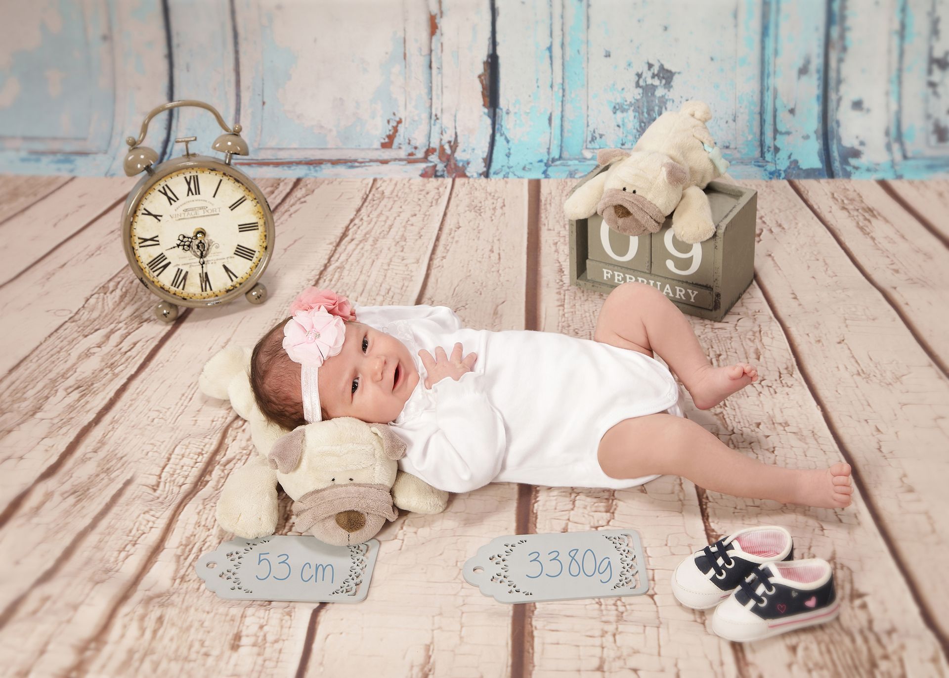 Baby liegt auf dem Boden in einer Deko-Szene mit Uhr, Stofftier und pastellfarbenem Hintergrund.
