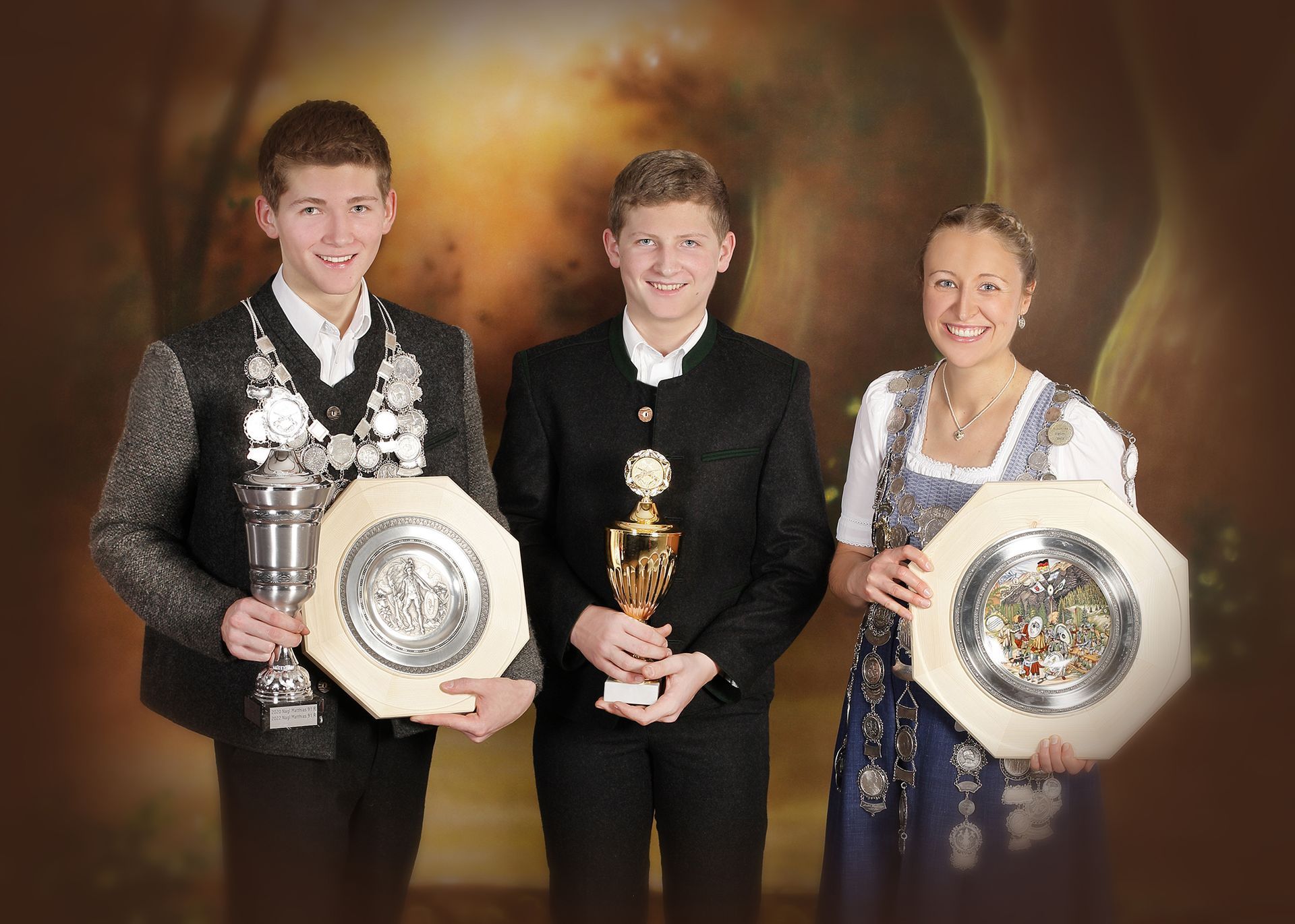 Drei Jugendliche in Tracht präsentieren stolz Pokale und Auszeichnungen.