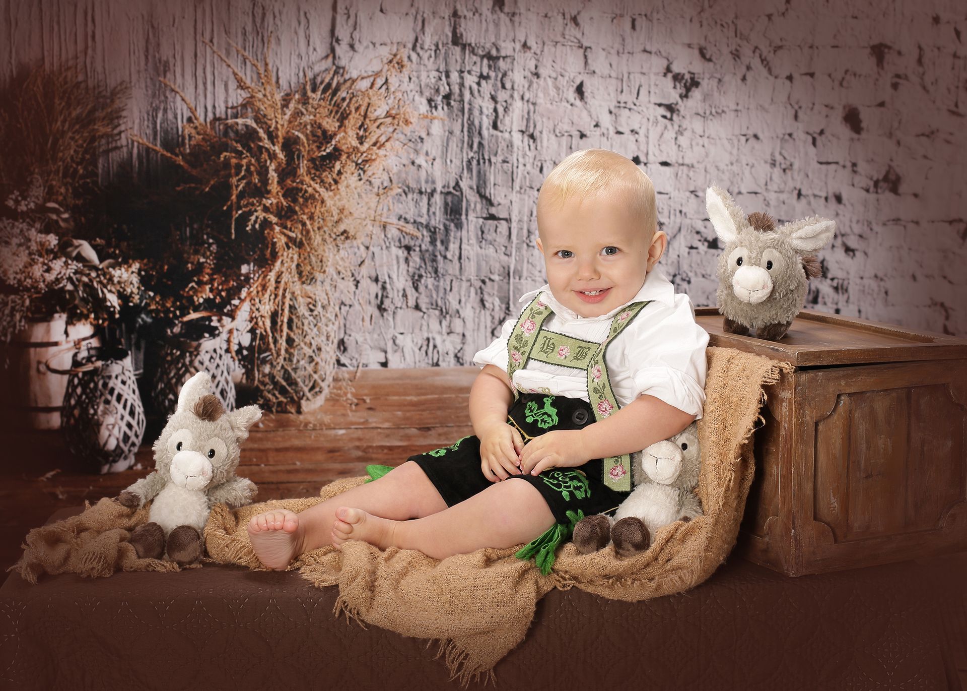 Lächelndes Kleinkind in Trachtenkleidung sitzt auf einem Tuch, umgeben von Stofftieren, vor rustikalem Hintergrund