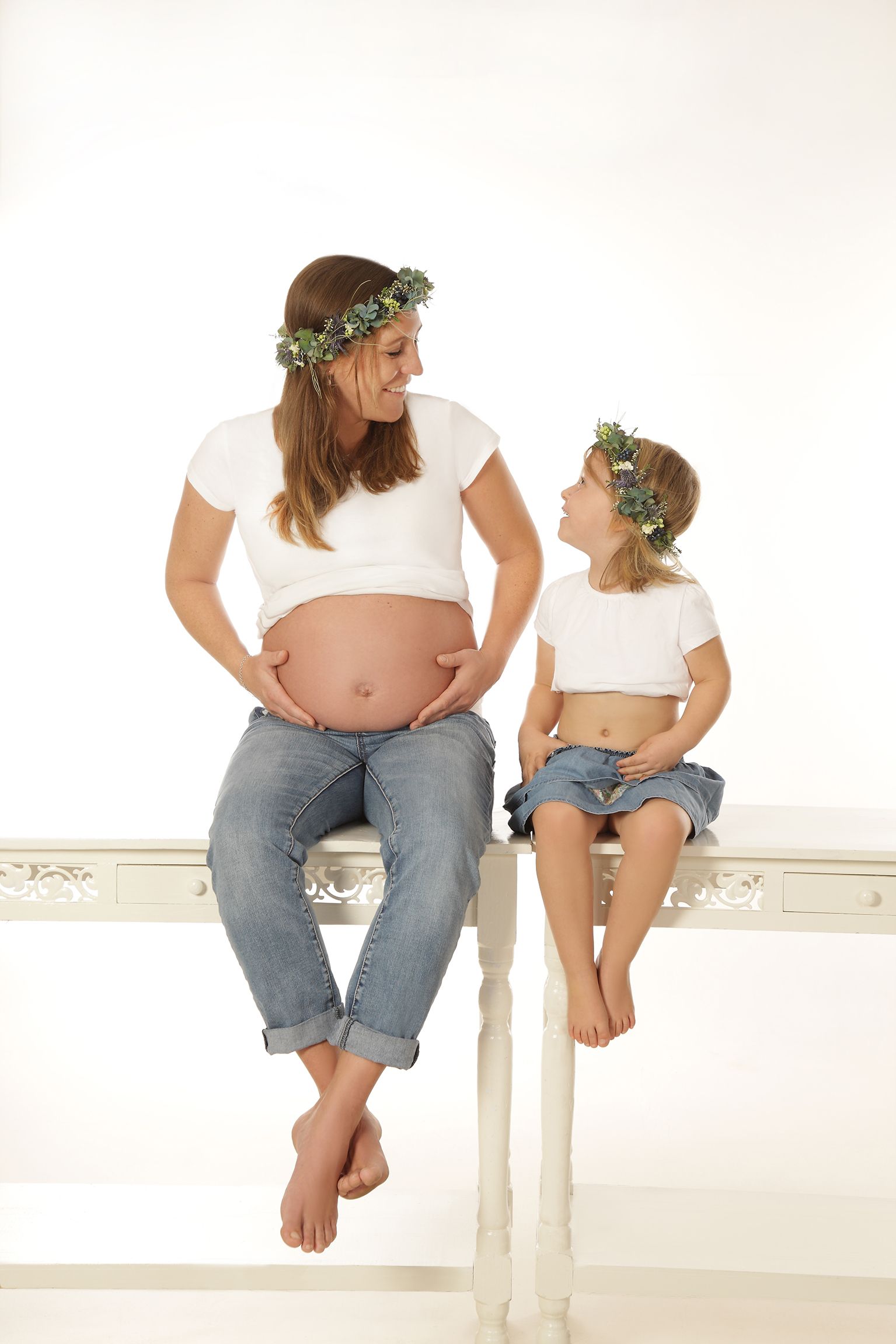Mutter und Kind gegenüber sitzend, beide mit Blumenkränzen auf dem Kopf und lächelnd.