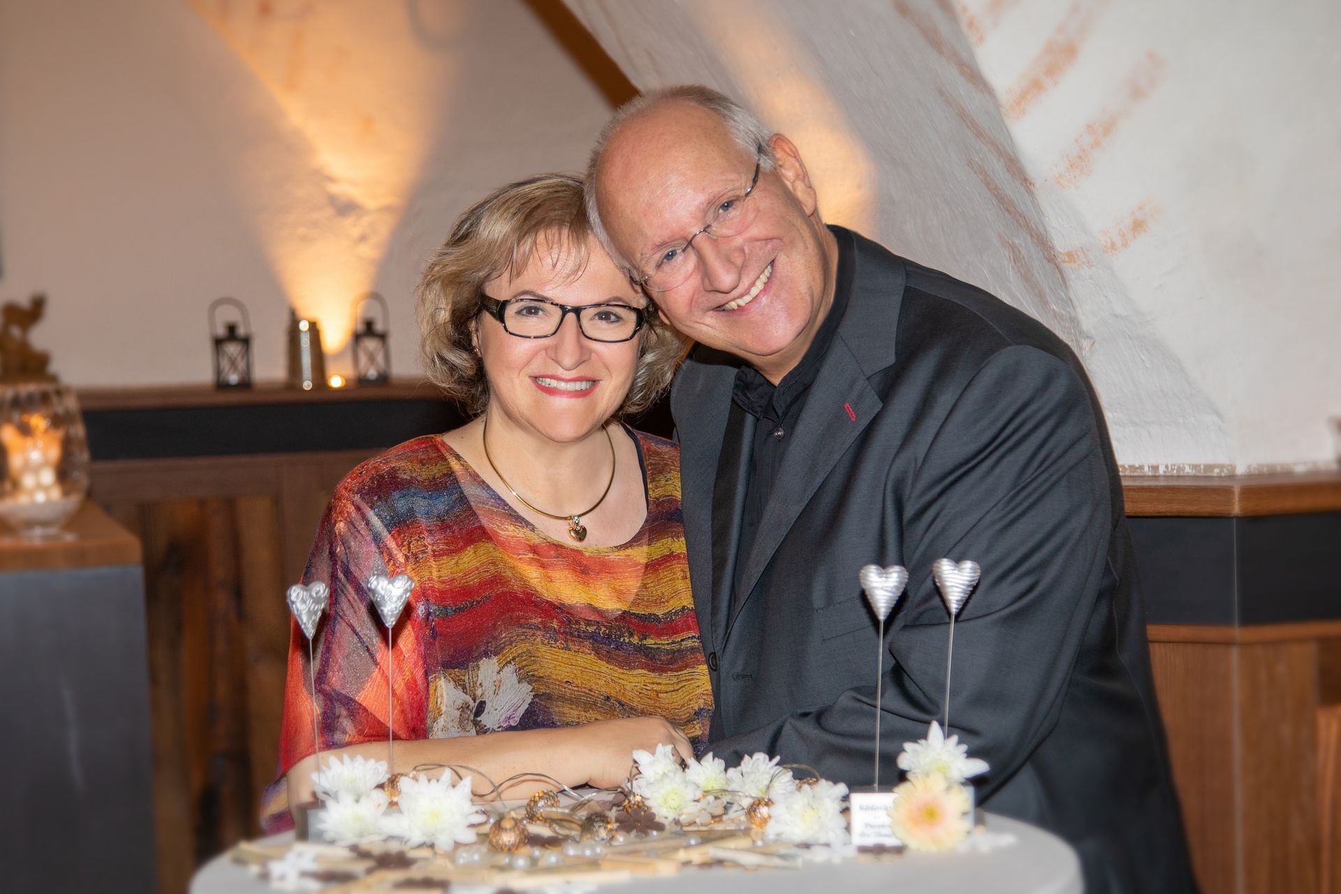 Porträt eines Paares, das lächelnd an einem Tisch sitzt, in warm beleuchtetem Ambiente.