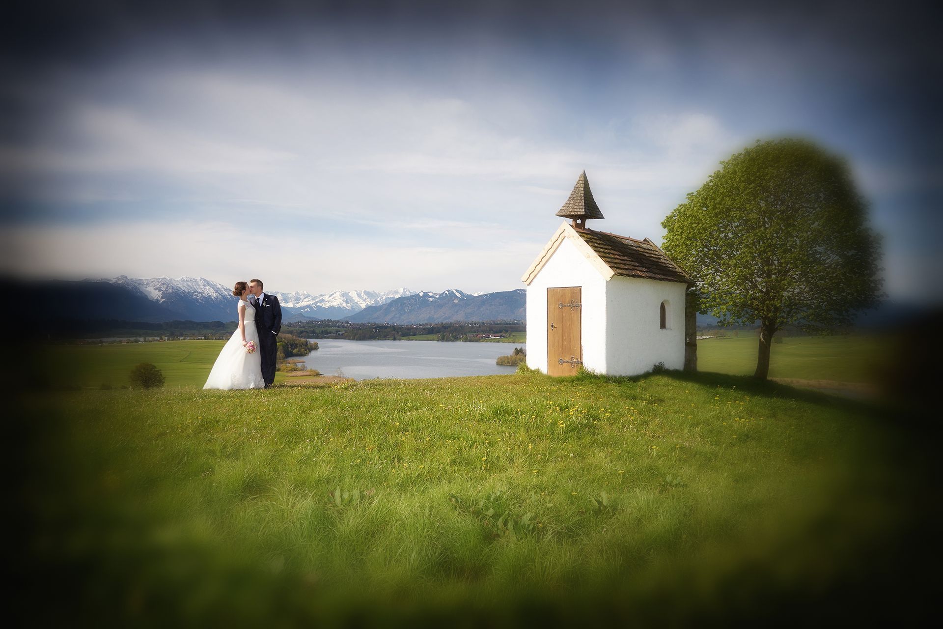 Hochzeitspaar steht vor einer kleinen Kapelle am Wasser mit Bergen im Hintergrund.