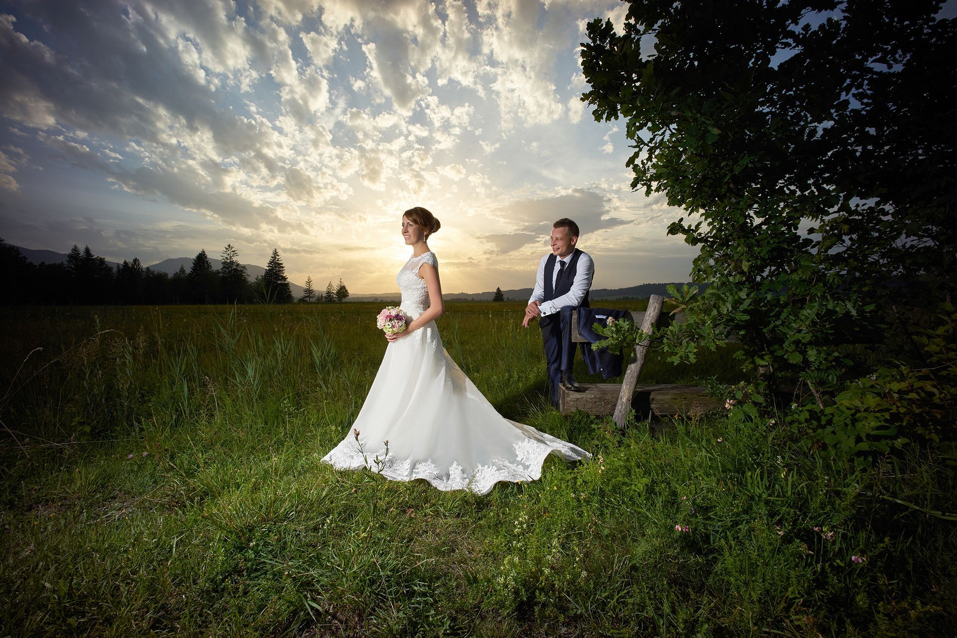 Braut und Bräutigam sitzen auf einer Bank im Grünen vor dramatischem Abendhimmel.