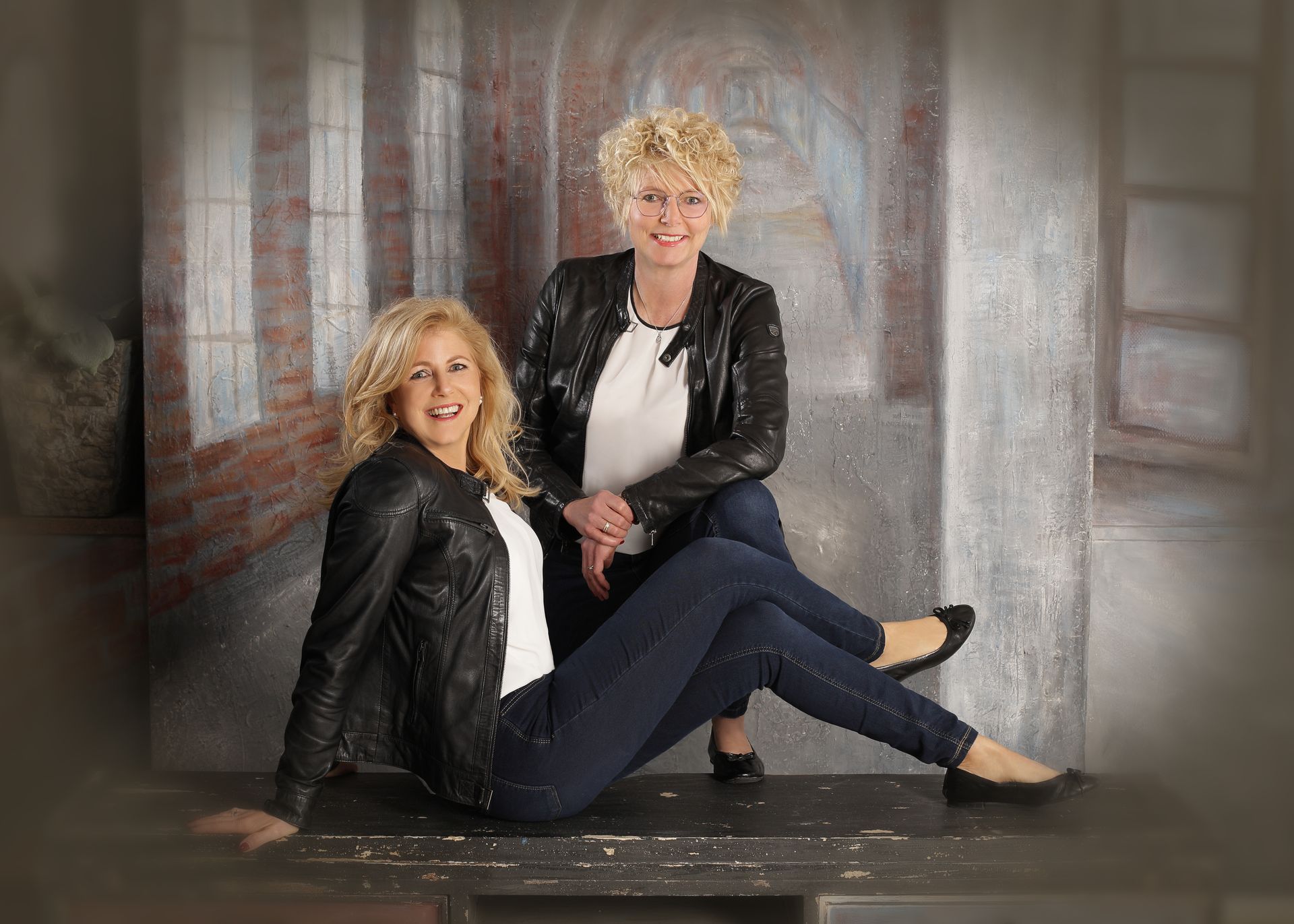 Zwei Frauen posieren lächelnd in einem Studio vor einem gemalten urbanen Hintergrund.