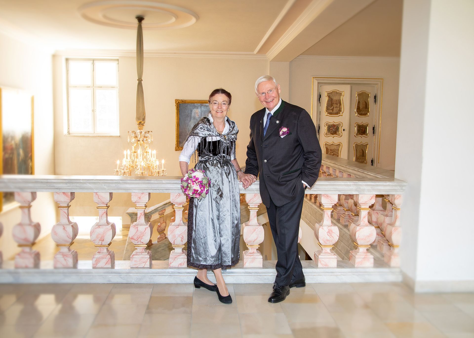 Elegantes Paar in festlicher Kleidung posiert in einem hellen Innenraum mit dekorativem Geländer.