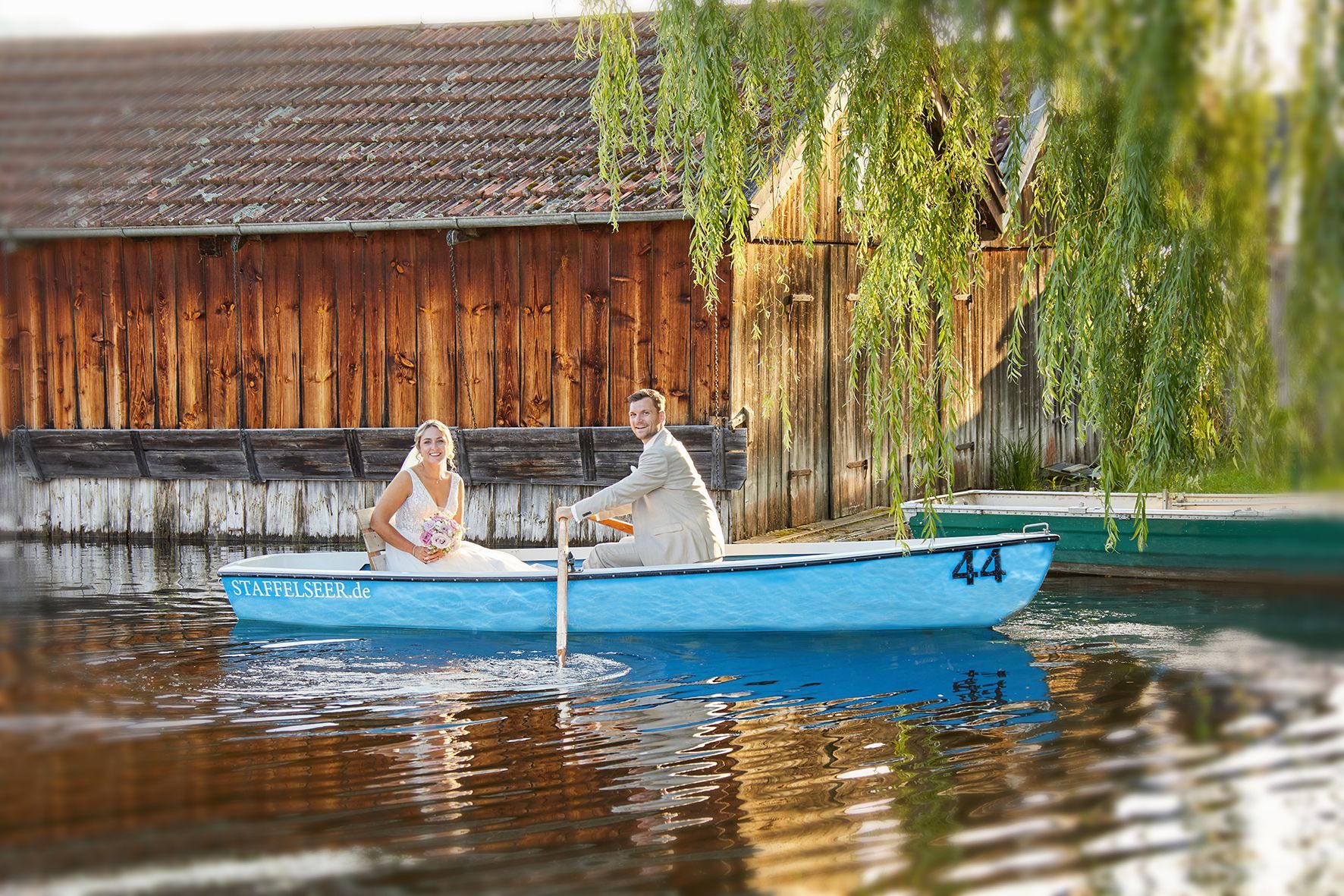 Paar sitzt in einem blauen Ruderboot auf einem See vor einer Holzscheune.