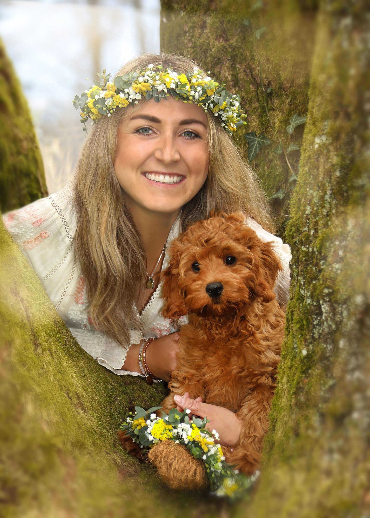 Frau mit Blumenkranz lächelt im Freien vor einem Baumstamm.
