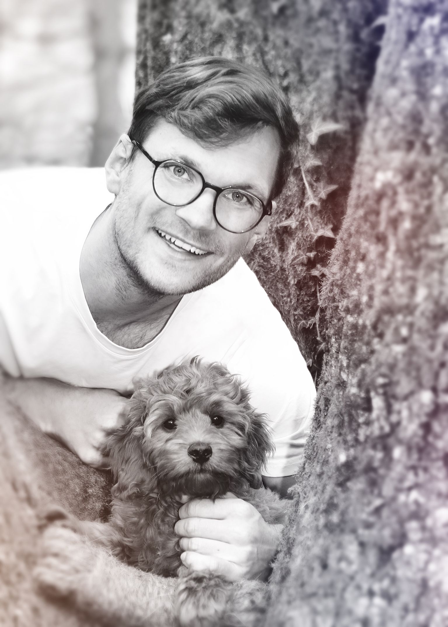 Schwarz-weiß-Porträt eines Mannes mit Brille, der lächelnd an einem Baum lehnt.