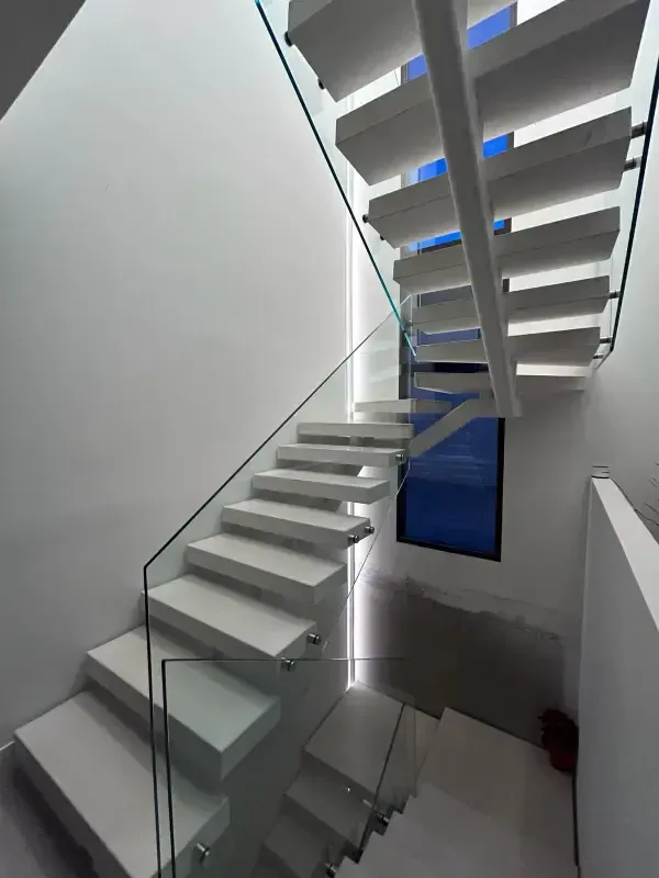Una escalera moderna de color blanco con barandillas de cristal asciende junto a una ventana alta y estrecha