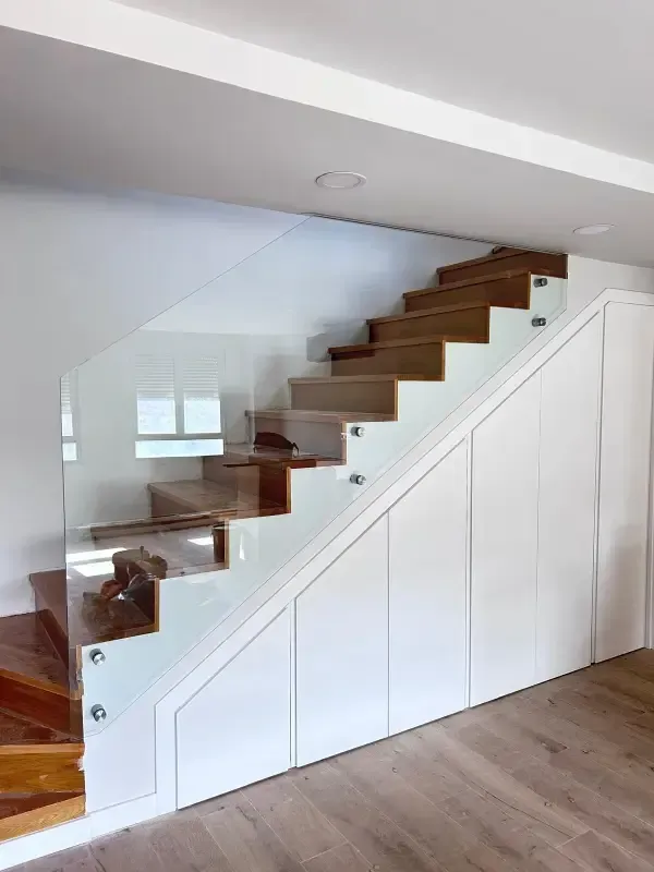 Una moderna escalera de madera con barandilla de cristal transparente y armarios empotrados blancos debajo.