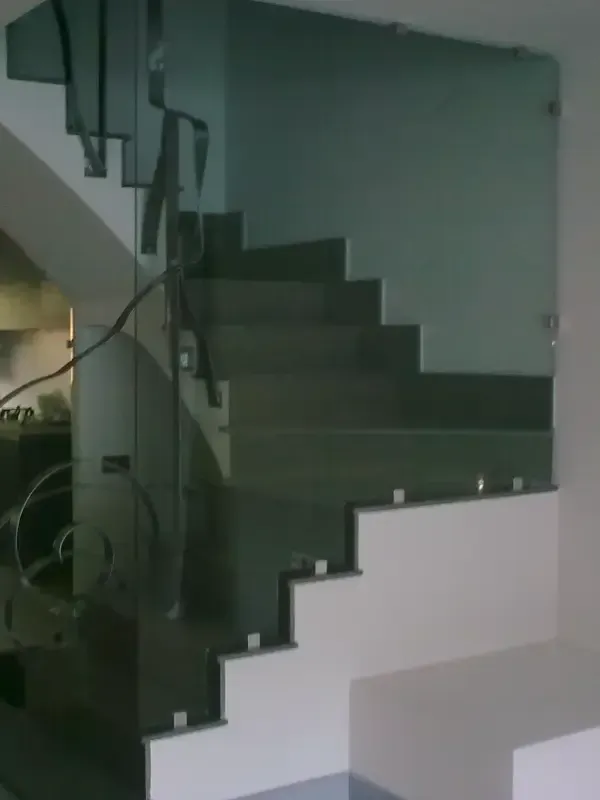 Escalera con peldaños oscuros y base blanca, con barandilla de cristal transparente sujeta con fijaciones metálicas.