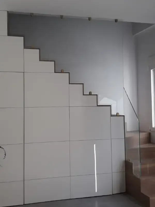 Un armario empotrado blanco con diseño escalonado sigue el perfil de una escalera junto a una barandilla de cristal.